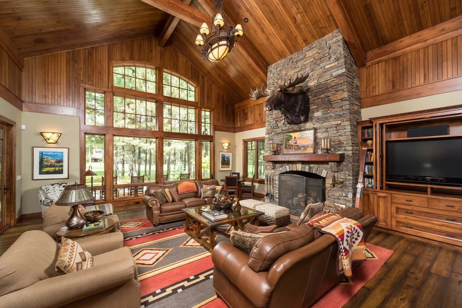 This living room, a highlight of a luxury custom home in Black Butte Ranch - Foin Follette, showcases masterful interior design by Kristy Yozamp. The space, with its wood-paneled walls, large windows, and a stone fireplace, creates a cozy retreat. A
