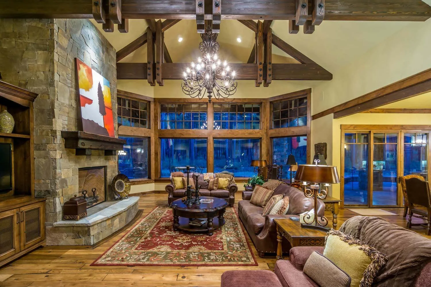 This living room, a key feature of a luxury custom home in Caldera Springs - Trailmere Circle, showcases masterful interior design by Kristy Yozamp. The space, with its large bay window, wood beams, and a grand stone fireplace, creates a cozy and ele