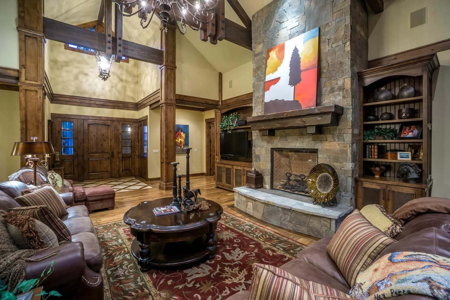 This cozy living room, a key feature of a luxury custom home in Caldera Springs - Trailmere Circle, showcases masterful interior design by Kristy Yozamp. The space, with its high ceiling, wooden beams, and a large stone fireplace, creates a warm and