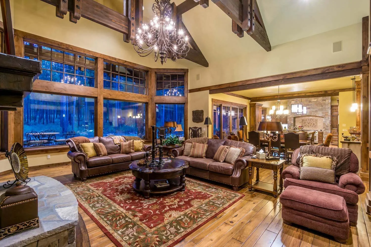This living room, a key feature of a luxury custom home in Caldera Springs - Trailmere Circle, showcases masterful interior design by Kristy Yozamp. The space, with its large windows, wooden beams, and a grand stone fireplace wall, is a beautiful exa