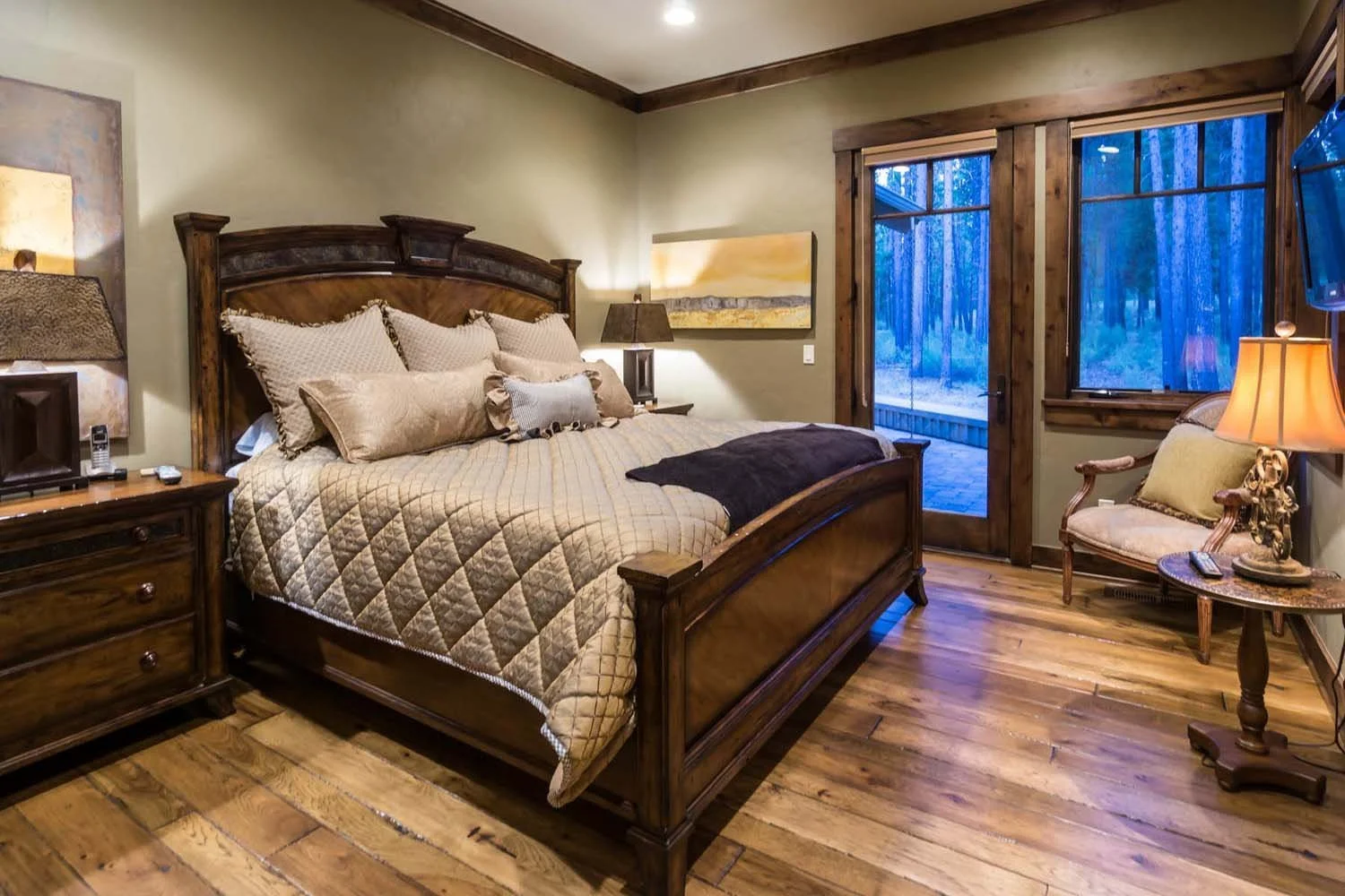This beautiful bedroom is a testament to the masterful architecture design by Spencer Williams. The space, with its elegant wooden bed and comfortable sitting chair, creates a cozy retreat in this Central Oregon home. The large windows and a door lea