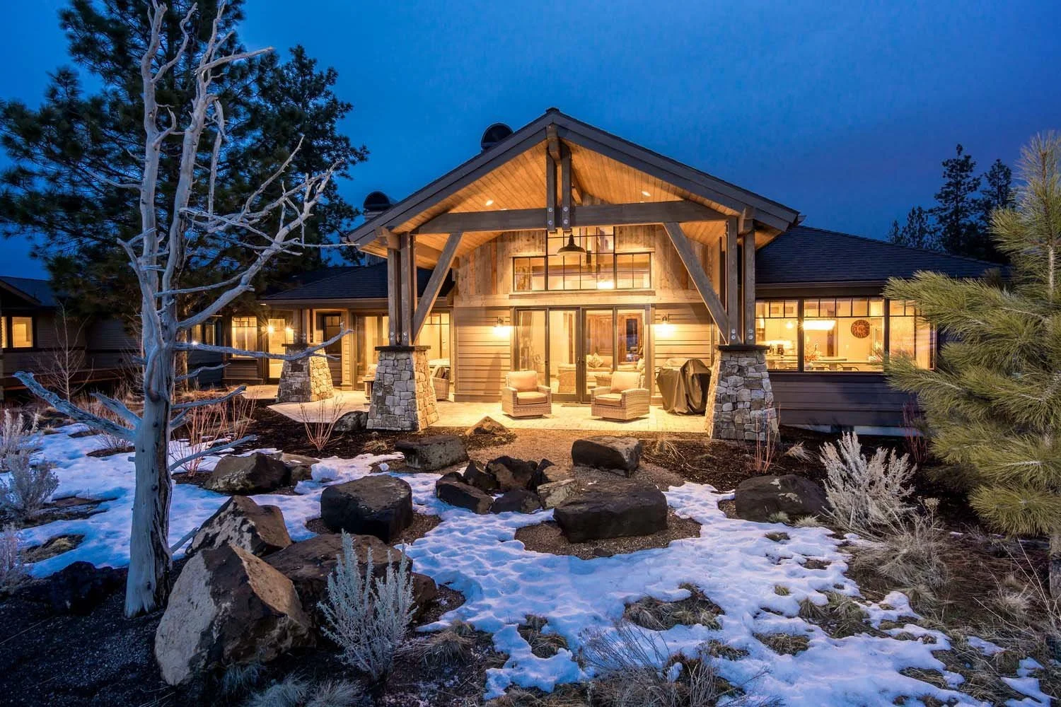 This luxury custom home in Tetherow - Hosmer Lake Drive is a masterpiece of architecture design by Spencer Williams. The modern house features a large, covered porch that is beautifully illuminated at night, creating a welcoming atmosphere. Surrounde