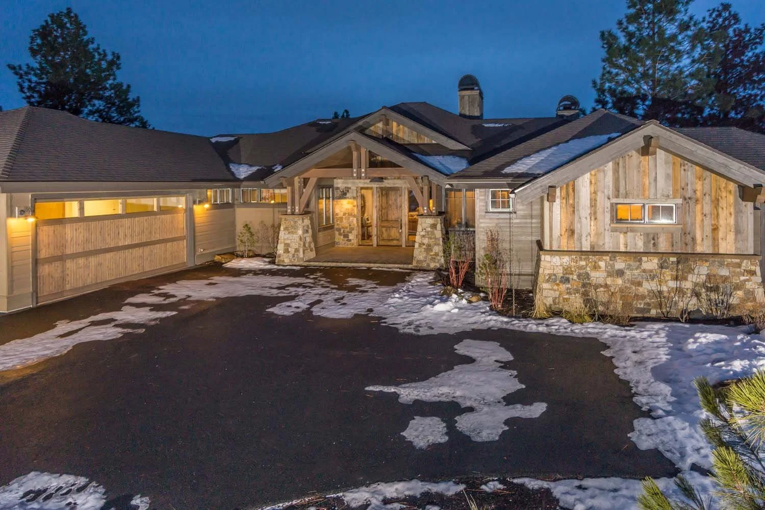 This luxury custom home in Tetherow - Hosmer Lake Drive showcases masterful architecture design by Spencer Williams. The house features a rustic exterior of wood and stone. The exterior, with its barnwood, natural stone, and heavy timber trusses, giv