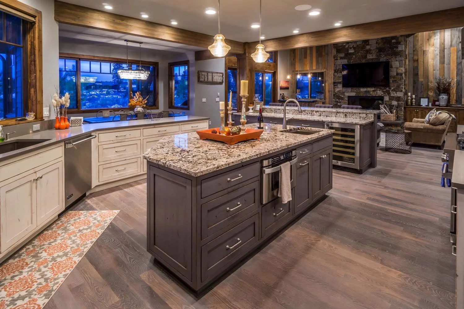 A spacious kitchen with a central dark gray island with a granite countertop, surrounded by white and gray cabinets. The kitchen features wooden flooring, large windows with dark wood frames, and multiple pendant lights hanging over the island. In th