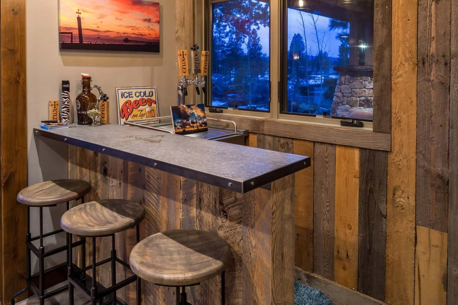 This rustic home bar, a key feature of a luxury custom home in Tetherow - Hosmer Lake Drive, is a testament to the masterful interior design by Kelly Warner. The space features three wood and metal bar stools, a gray countertop, and beer taps, creati