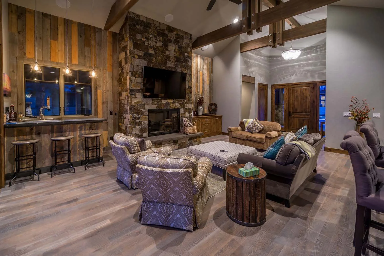 This cozy living room in Tetherow - Hosmer Lake Drive is a showcase of masterful interior design by Kelly Warner. The space, which has the feel of a mountain lodge, features a stone fireplace, a wall-mounted TV, and multiple sofas and armchairs. The