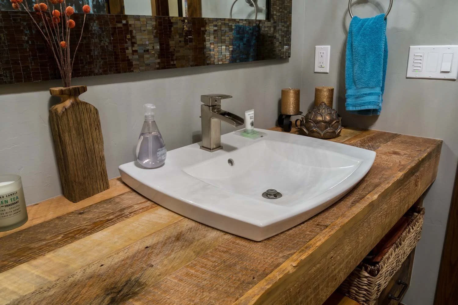This bathroom vanity, a key feature of a luxury custom home in Tetherow - Hosmer Lake Drive, showcases masterful interior design by Kelly Warner. The space features a wooden countertop with a wooden vase, a soap dispenser, and gold candles, reflectin