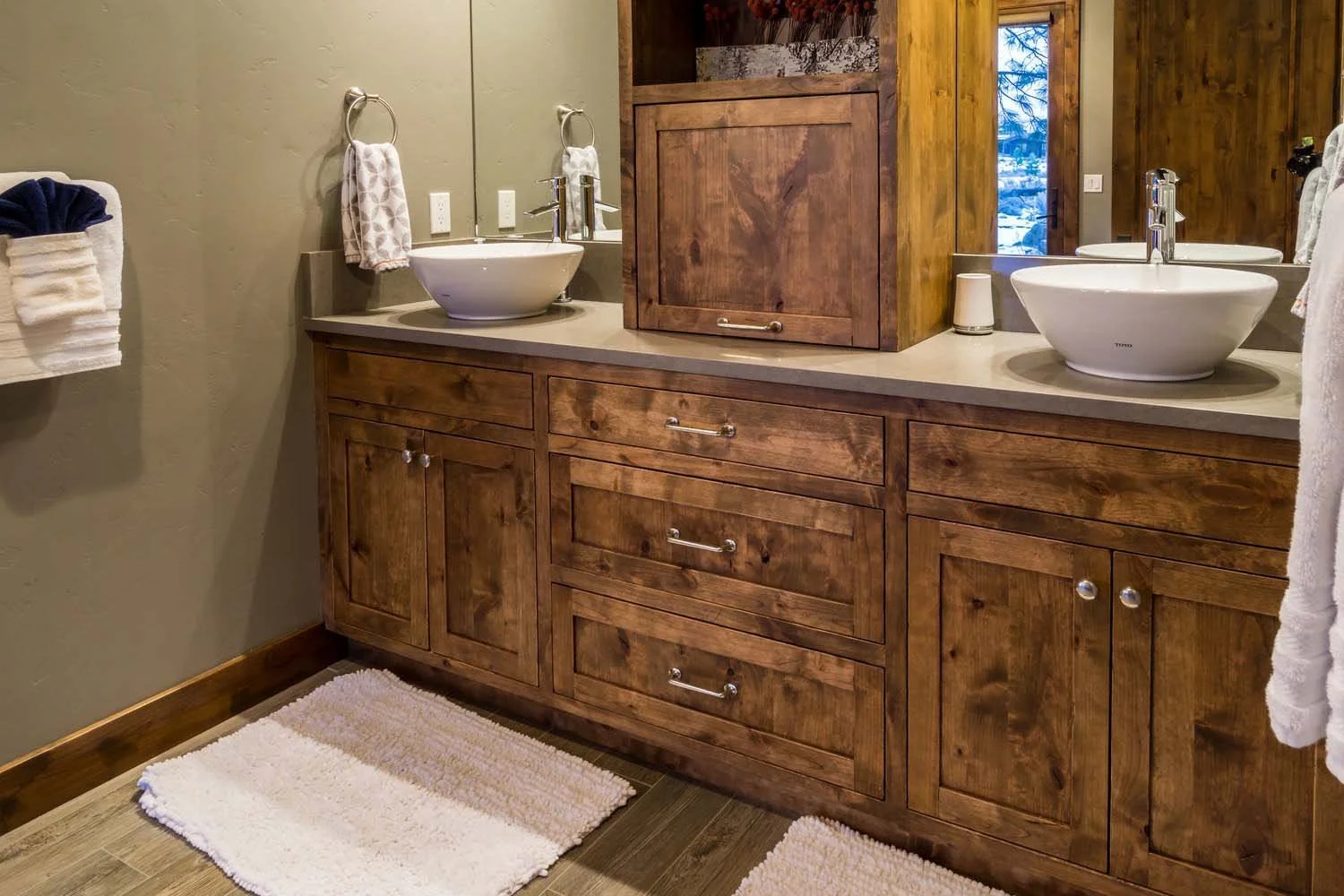 This rustic bathroom in Tetherow - Hosmer Lake Drive, a luxury custom home, is a testament to the masterful interior design by Kelly Warner. The space features a double vanity with wooden cabinets and a beige countertop, two oval vessel sinks with mo