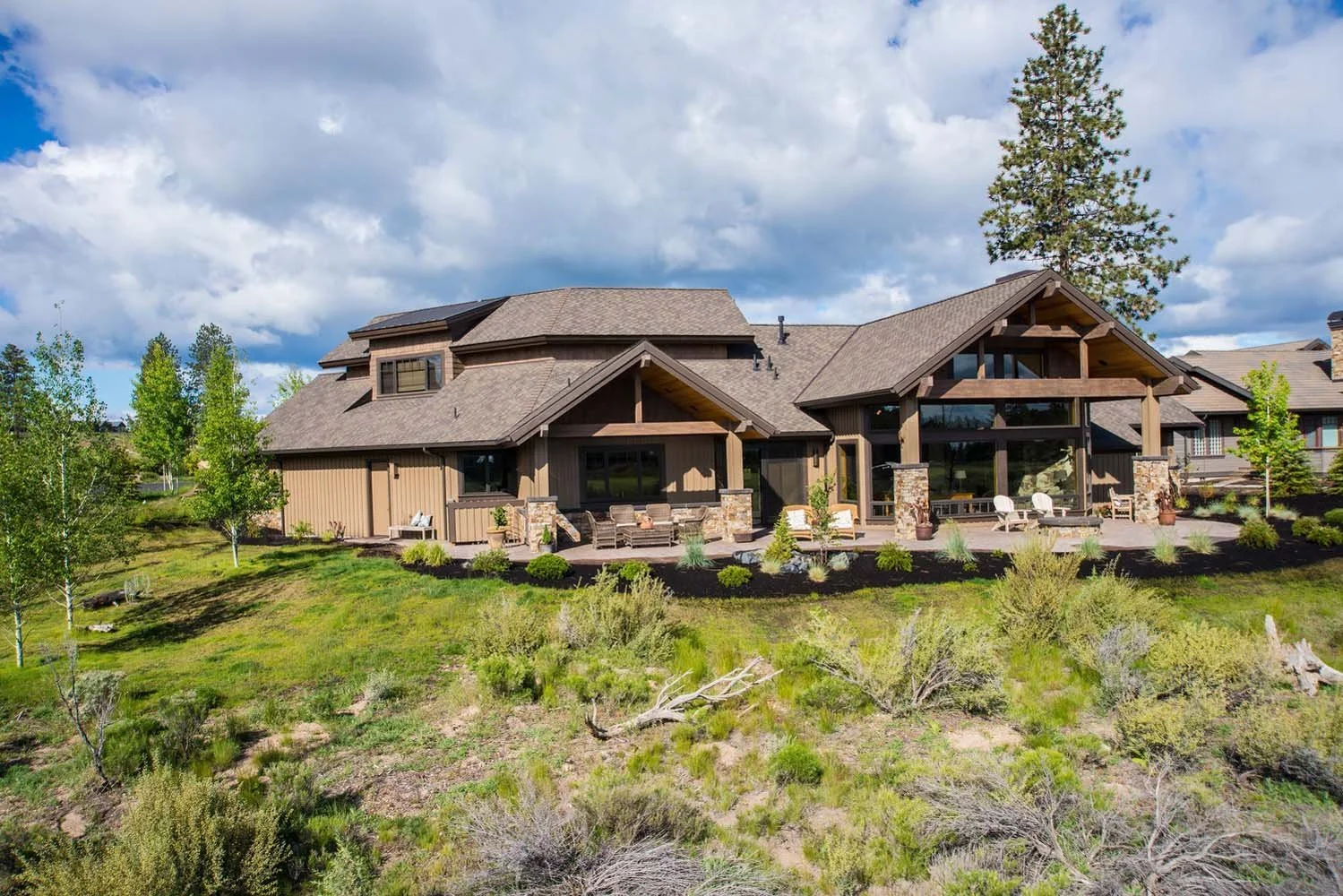 This luxury custom home in Tetherow - Weinhard Court, designed by Spencer Williams, is a masterpiece of modern architecture. The large house features beautiful wooden and stone accents and is surrounded by lush greenery. The spacious backyard with pa