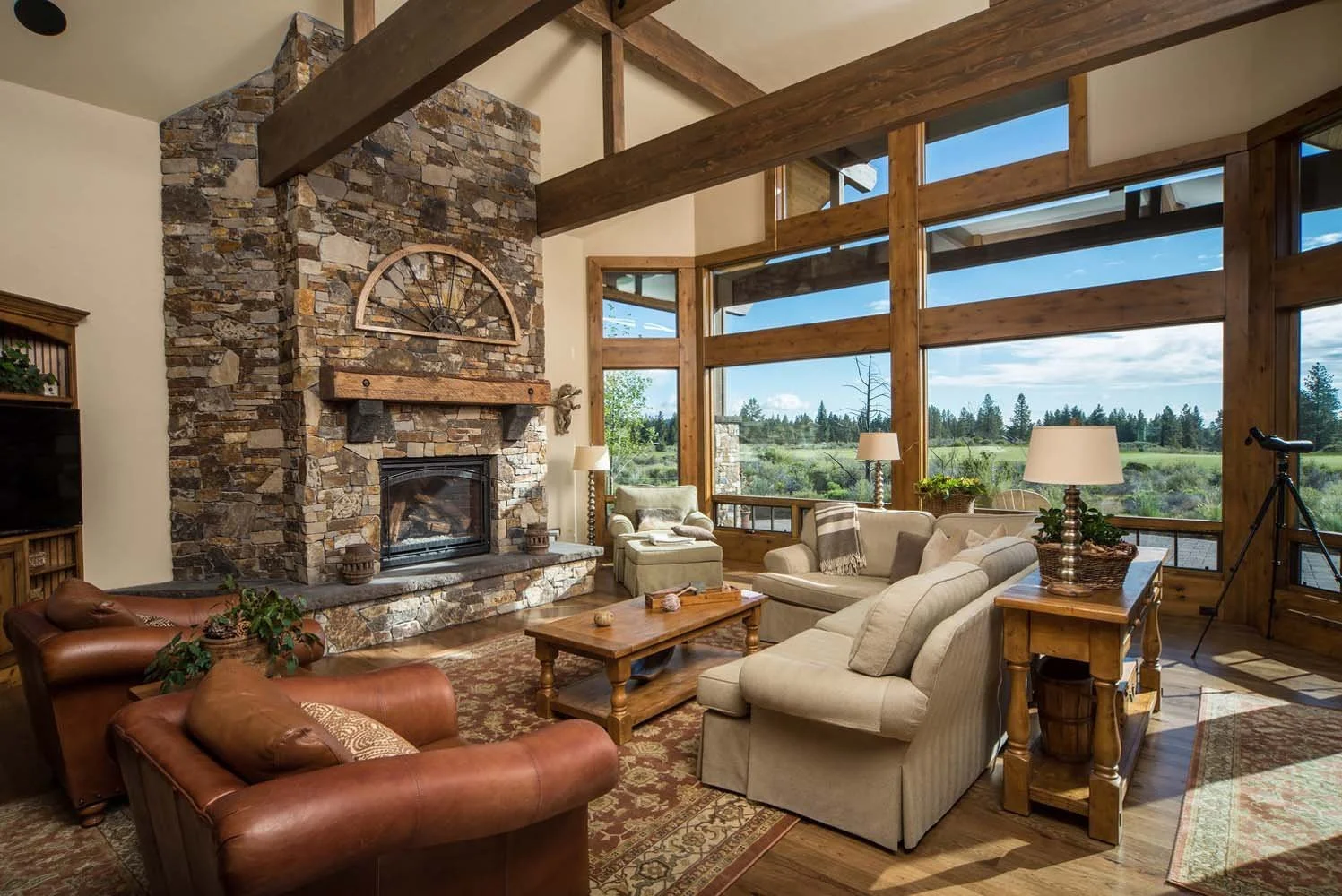 This living room, a key feature of a luxury custom home in Tetherow - Weinhard Court, is a showcase of masterful interior design by Kristy Yozamp. The space, with its large stone fireplace, cozy beige sofas, and a wooden coffee table, creates a warm