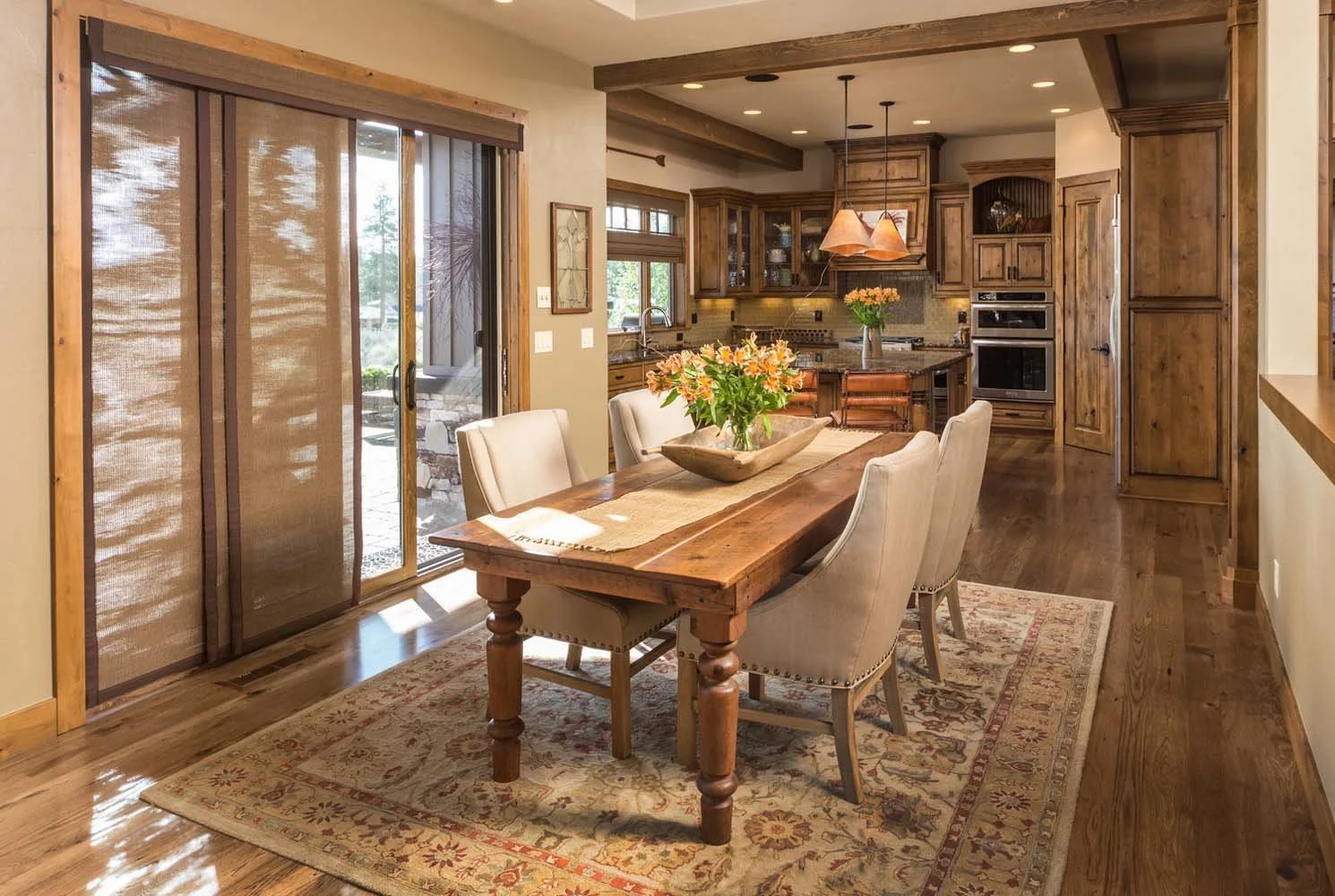 This elegant dining room and kitchen, a key feature of a luxury custom home in Tetherow - Weinhard Court, showcases masterful interior design by Kristy Yozamp. The space, with its wooden table and chairs, leads to a kitchen with wooden cabinetry and