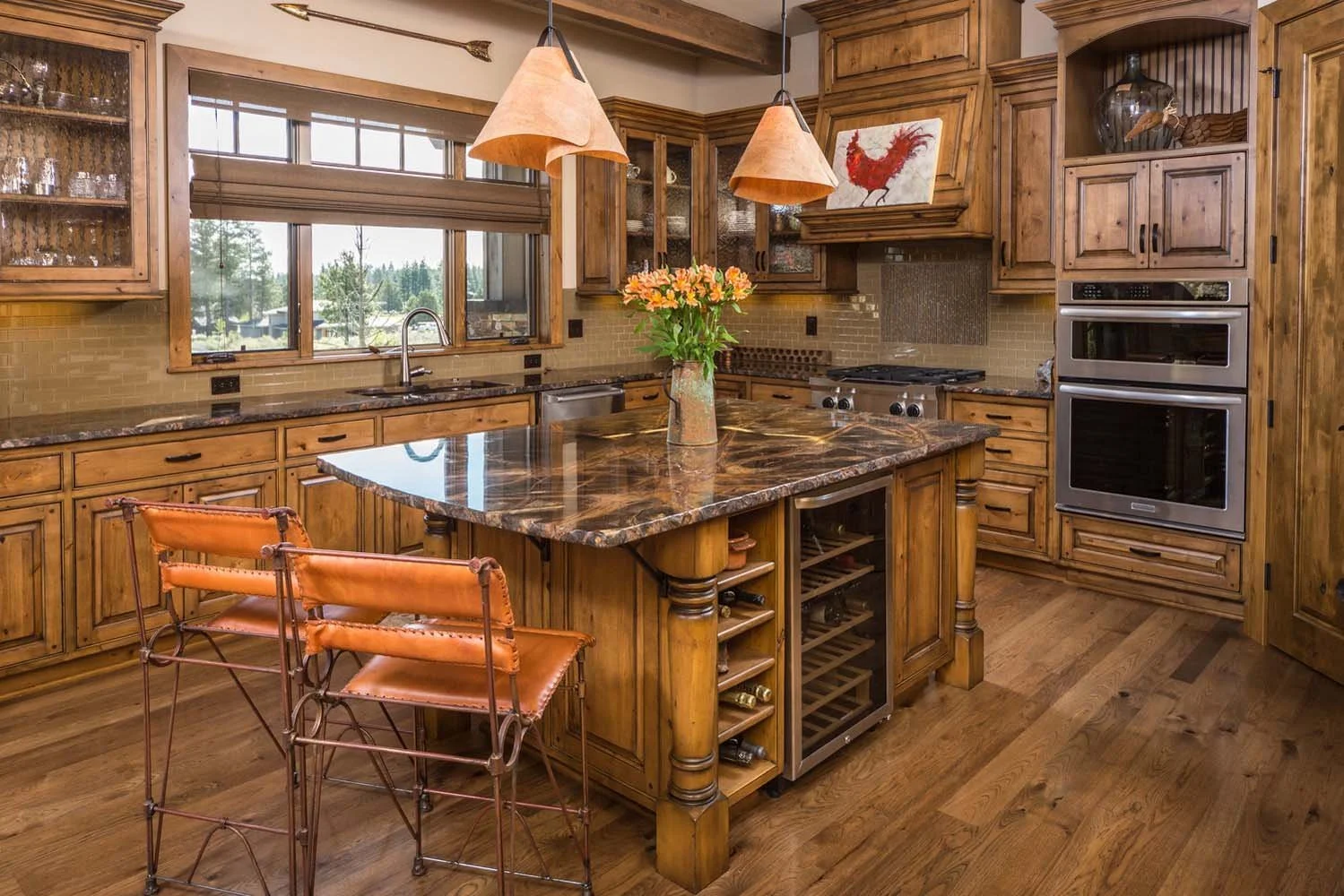 This rustic kitchen in Tetherow - Weinhard Court, a luxury custom home, showcases masterful interior design by Kristy Yozamp. The space features wooden cabinets, a marble countertop island with barstools, and modern pendant lights. The inclusion of d