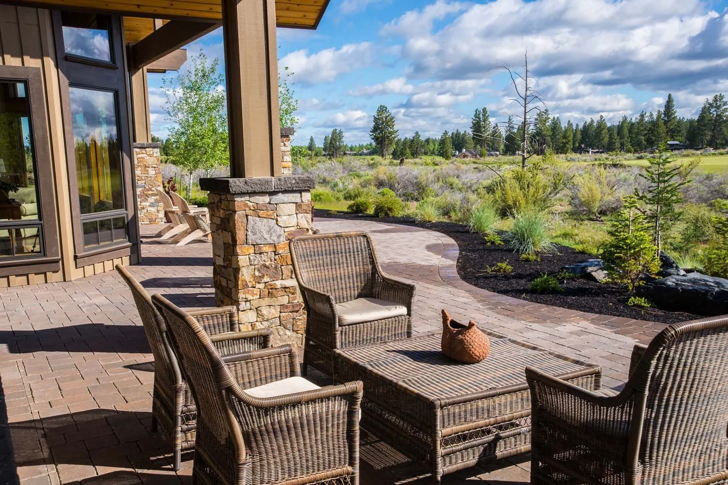 This outdoor patio, a key feature of a luxury custom home in Tetherow - Weinhard Court, showcases masterful interior design by Kristy Yozamp. The space, with its comfortable wicker chairs and a coffee table, overlooks a beautifully landscaped yard. T