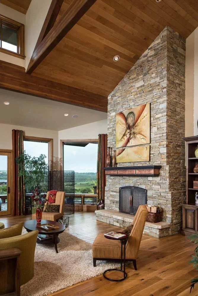 This superb living room, a highlight of a luxury custom home in Tetherow - Skene Trail, is a showcase of masterful interior design by Kristy Yozamp. The space, with its large stone fireplace and wooden ceiling, creates a cozy and elegant atmosphere.