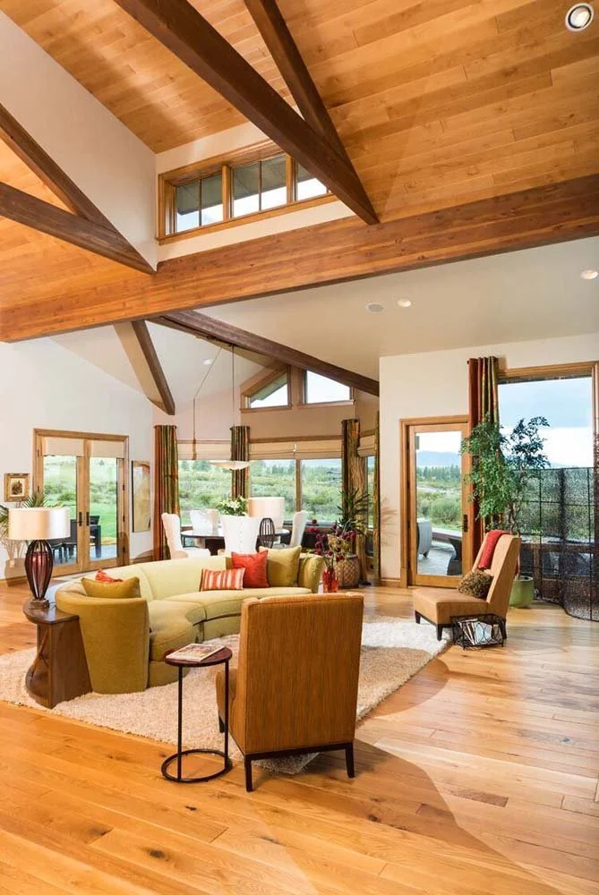 This spacious living room in Tetherow - Skene Trail, a luxury custom home, is a showcase of masterful interior design by Kristy Yozamp. The space, with its high wooden ceilings and large windows, is designed to bring in natural light and connect with