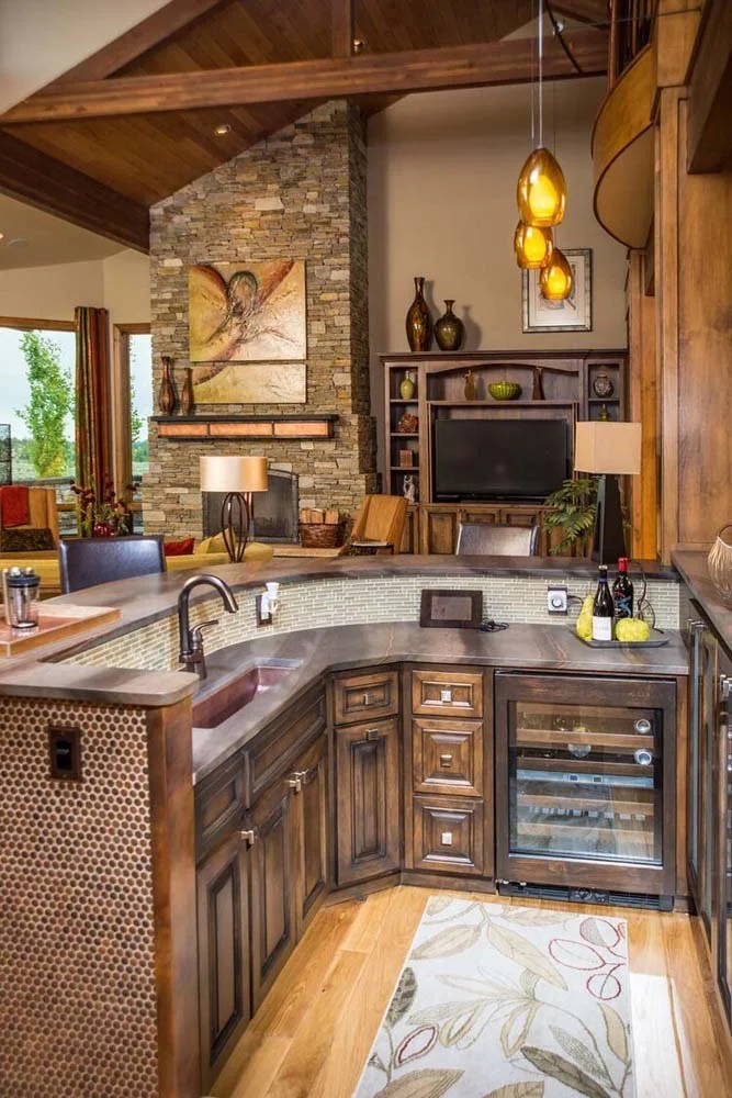 This modern kitchen in Tetherow - Skene Trail, a luxury custom home, is a beautiful showcase of interior design by Kristy Yozamp. The open-concept space, with its wooden cabinets and a stone fireplace, creates a warm and inviting atmosphere. The seam