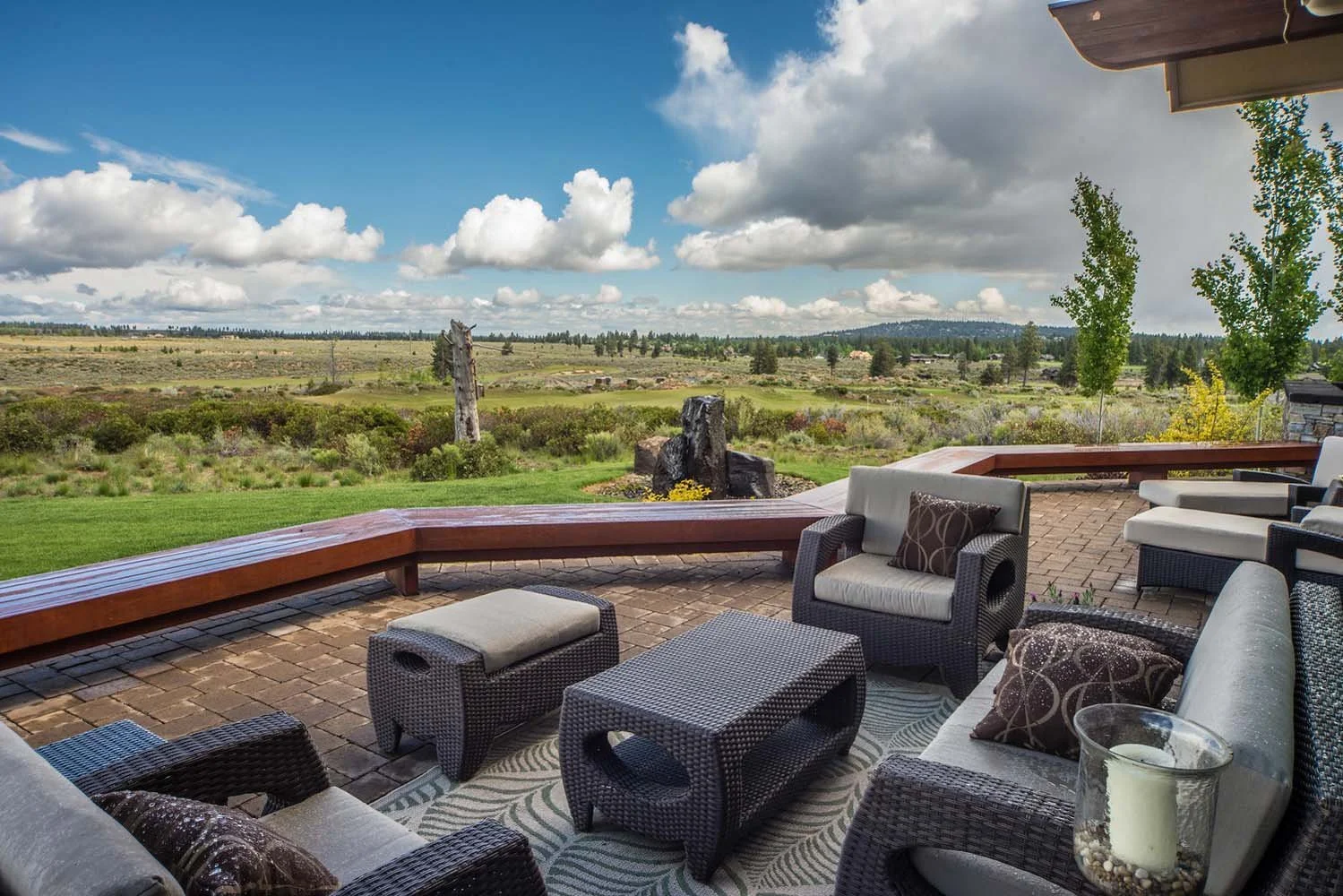 This modern outdoor patio, a key feature of a luxury custom home in Tetherow - Skene Trail, showcases the masterful interior design by Kristy Yozamp. The space, with its comfortable wicker chairs, a bench, and cushions, overlooks a lush green field,