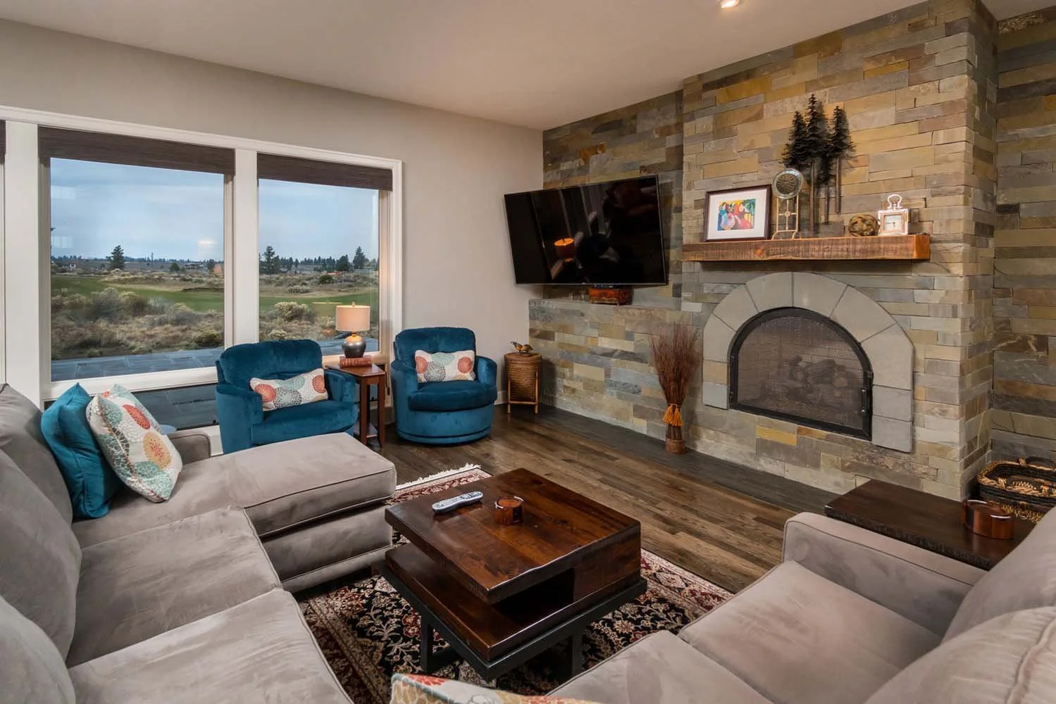 Explore the captivating architecture of this luxury custom home in Tetherow - Skene Trail. The living room features a stone fireplace, large windows with a scenic view, and comfortable sofas and armchairs. The thoughtful interior design by Kristy Yoz