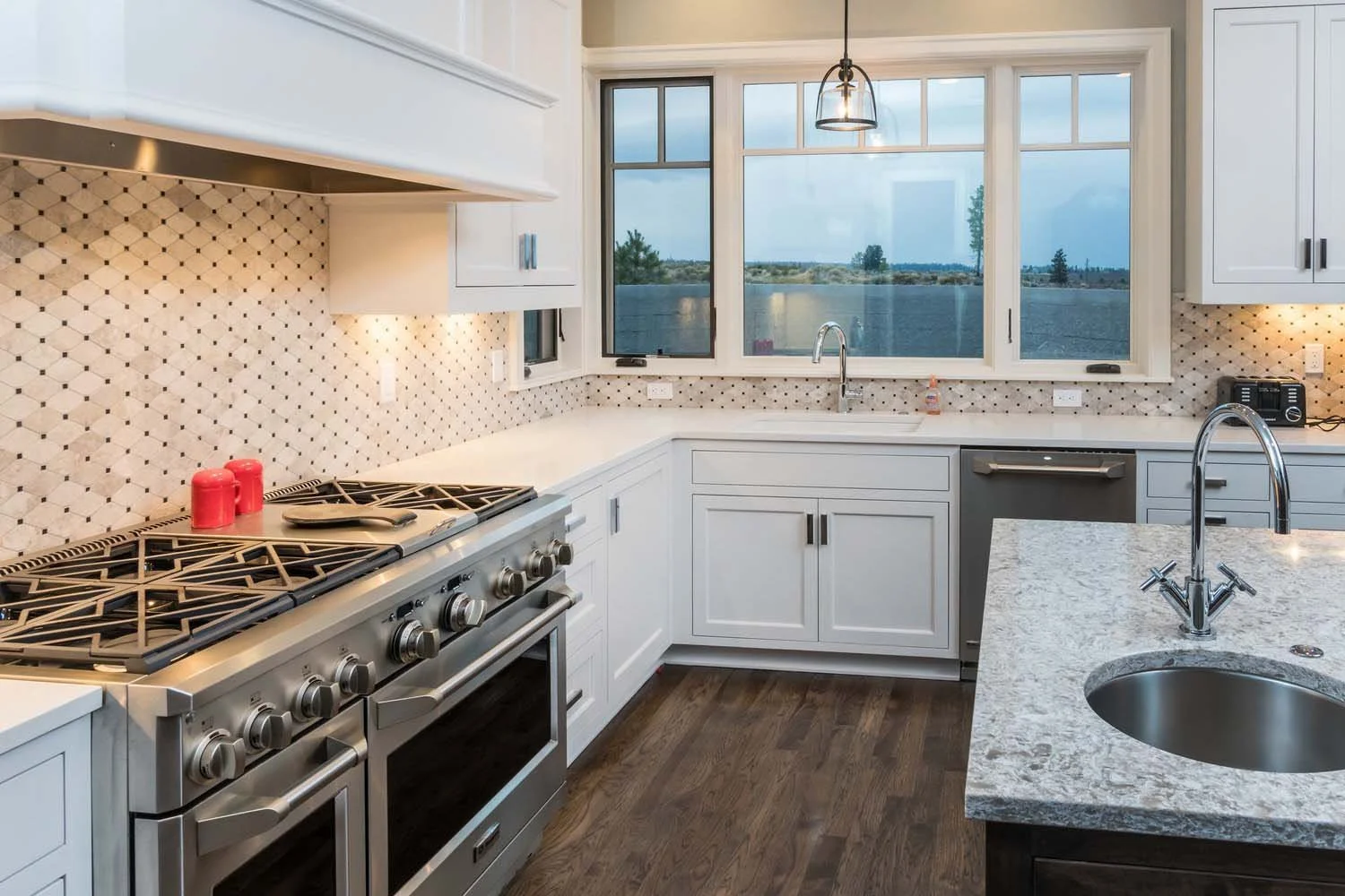 This modern kitchen in Tetherow - Skene Trail, a luxury custom home, is a beautiful showcase of interior design by Kristy Yozamp. The space, with its elegant white cabinets, a large island with a granite countertop, and a patterned tile backsplash, i