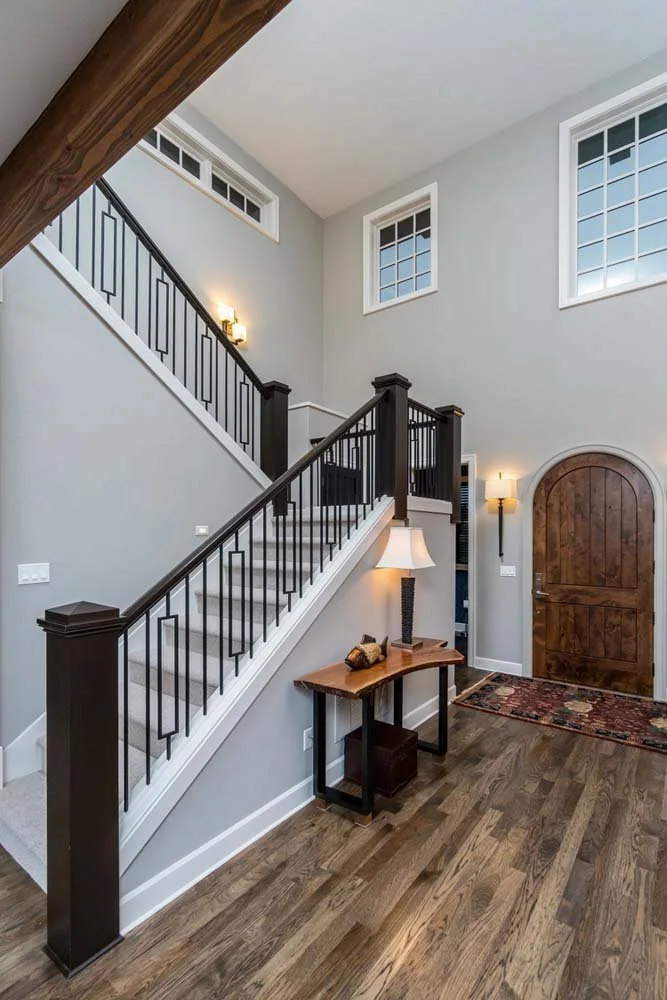 This home entryway in Tetherow - Skene Trail, a luxury custom home, is a beautiful showcase of interior design by Kristy Yozamp. The space, with its elegant wooden door, hardwood floors, and a staircase with a black railing, creates a welcoming and s