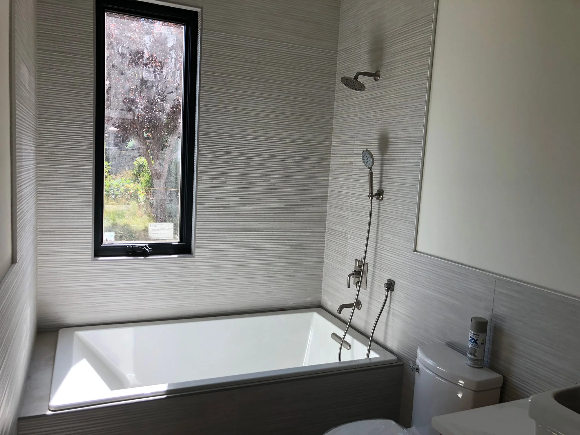 Modern bathroom with a white bathtub, a window showing trees outside, a showerhead with a handheld attachment, a toilet with a can of air freshener on top, and part of a sink.