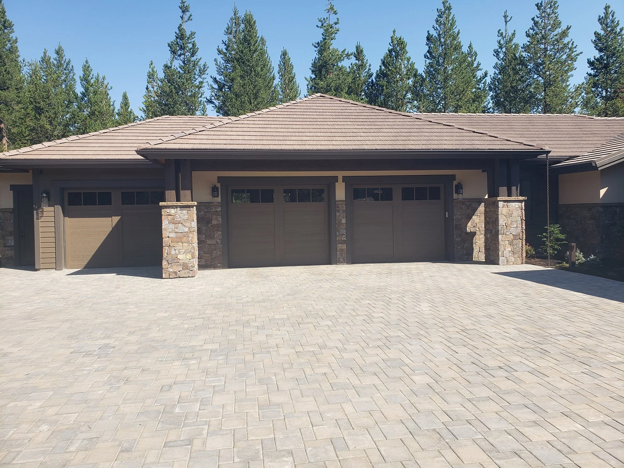 View of a modern house with a stone and wood exterior, three garage doors, and a tiled hip roof, surrounded by forest trees under a clear blue sky.