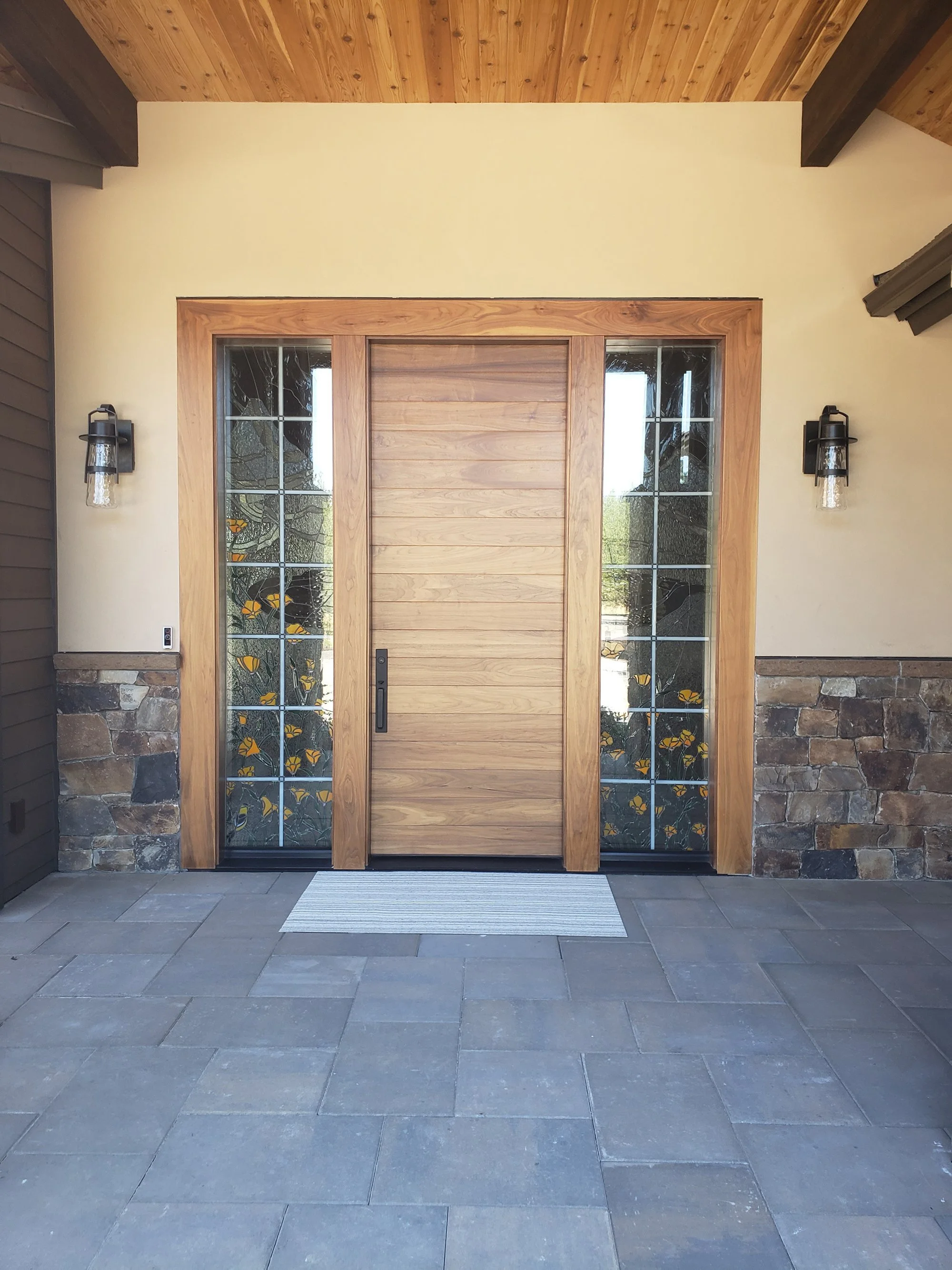 Front door with wooden frame, sidelight windows with butterfly designs, stone wall accents, and outdoor lighting fixtures.