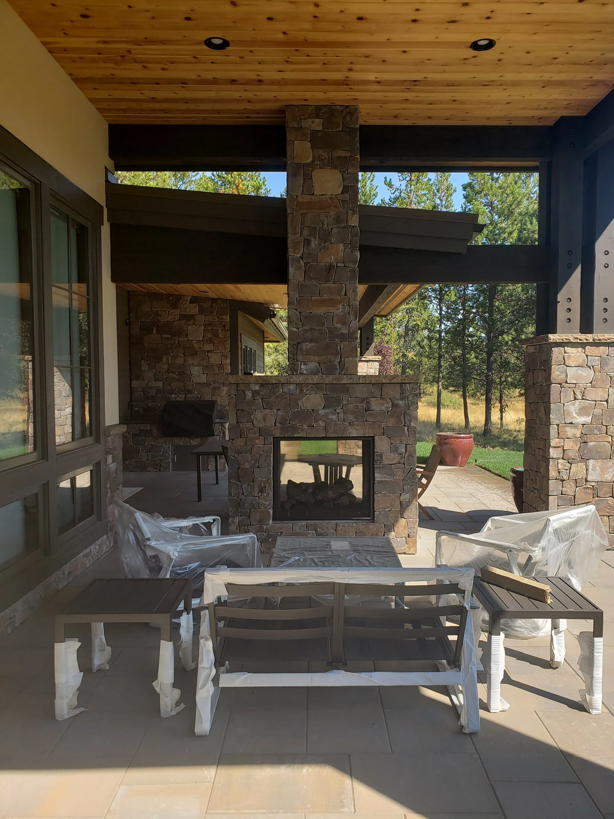 Covered patio with wooden ceiling, stone fireplace, and outdoor furniture wrapped in plastic.