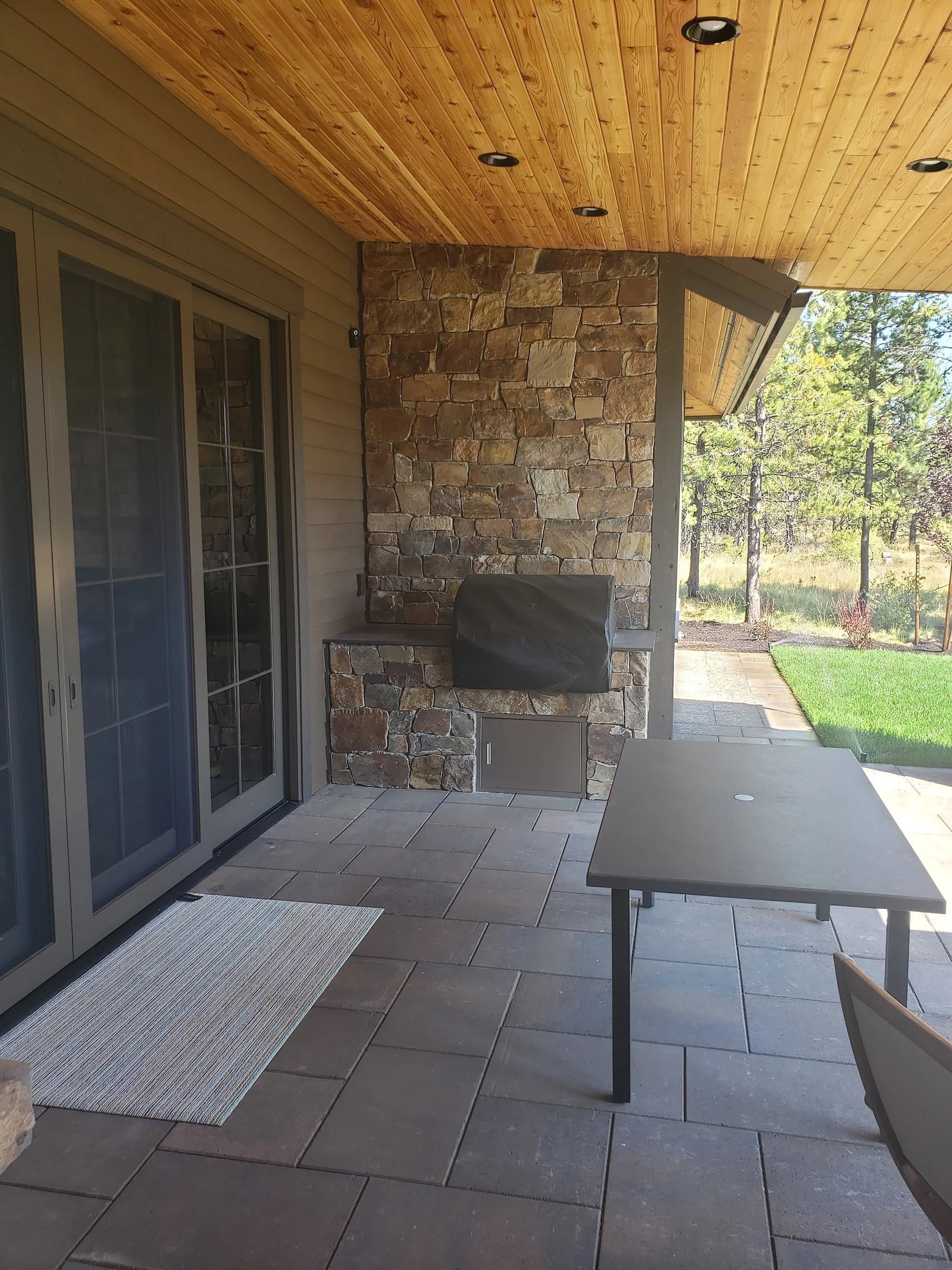 Outdoor patio area with stone fireplace, sliding glass door, small table, ceiling lights, and lawn with trees in background.