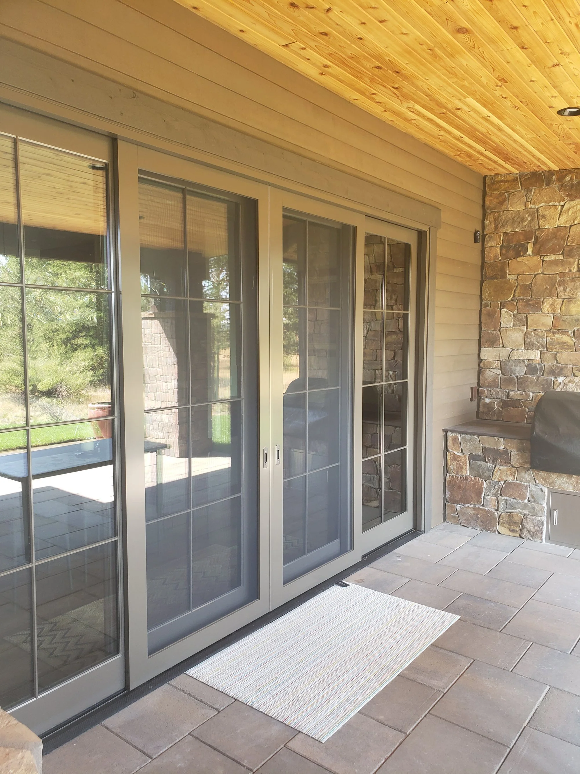 Sliding glass door on a patio with beige frames, a stone wall to the right, and a tiled floor, with a striped rug in front of the door.