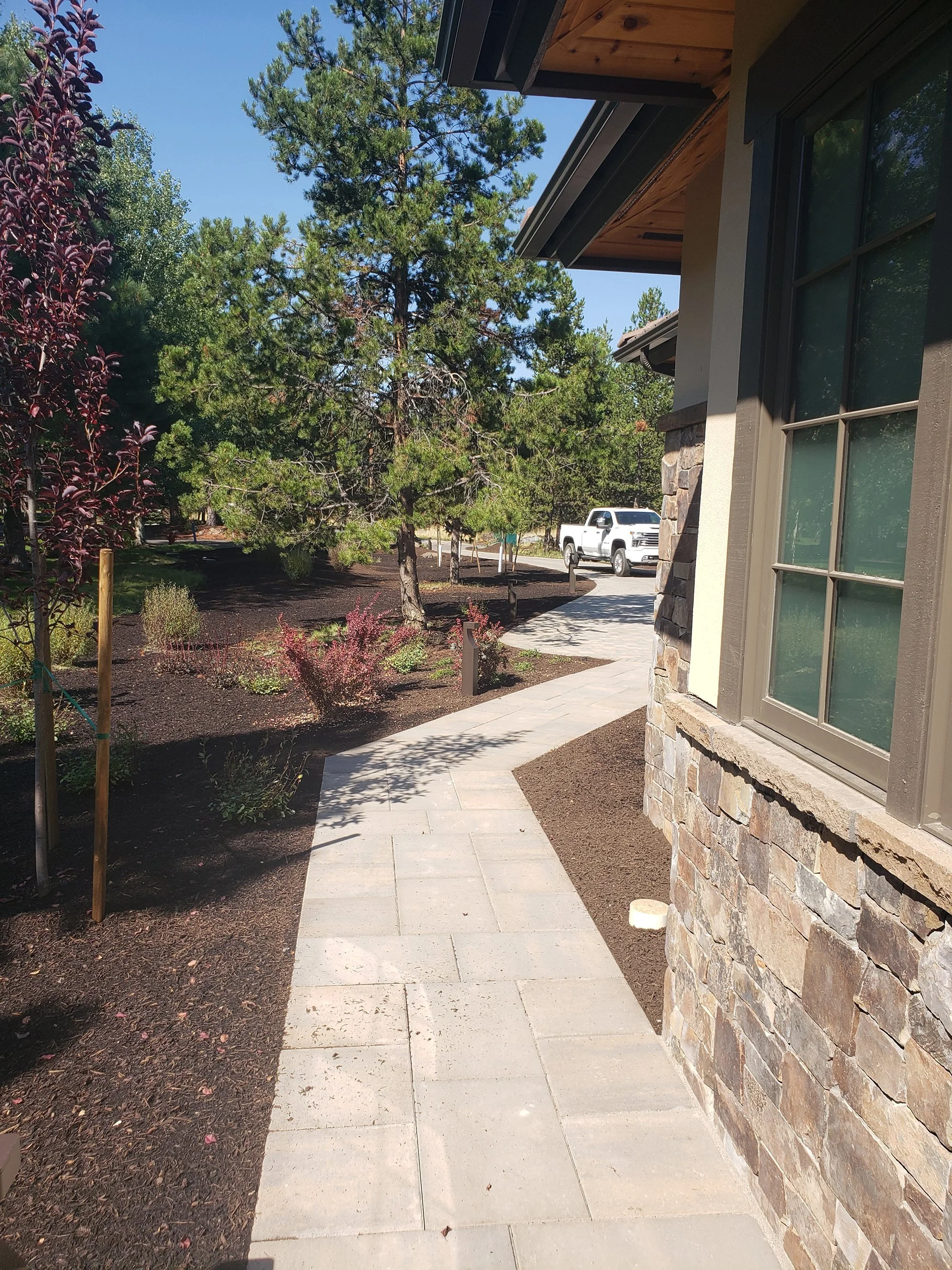 Sidewalk leading around a house with stone exterior walls, surrounded by freshly tilled soil and landscaping plants, with trees and a parked white pickup truck in the background on a sunny day.