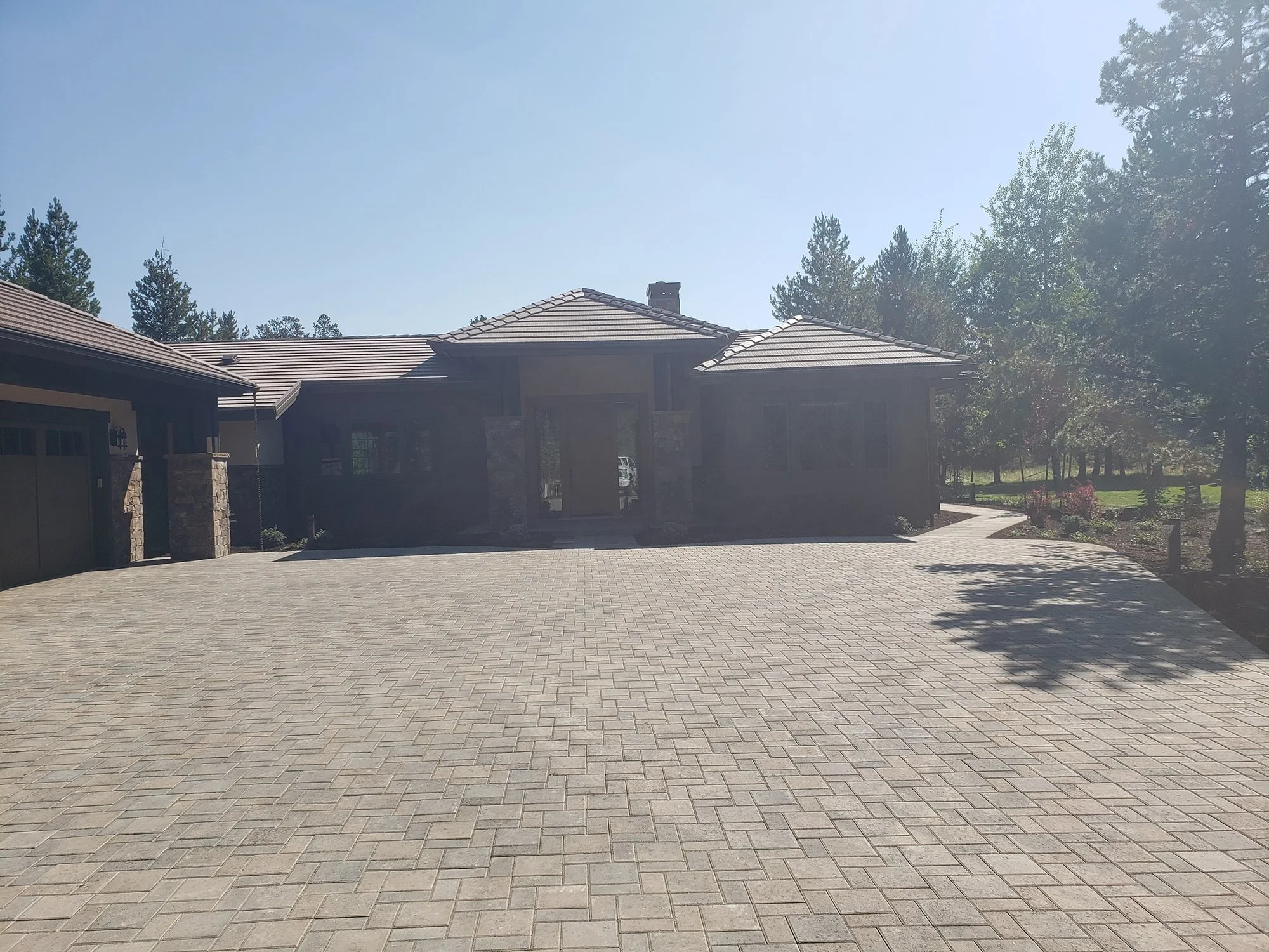 Large modern house with stone accents, tiled roof, and a spacious paved driveway, surrounded by trees and greenery.