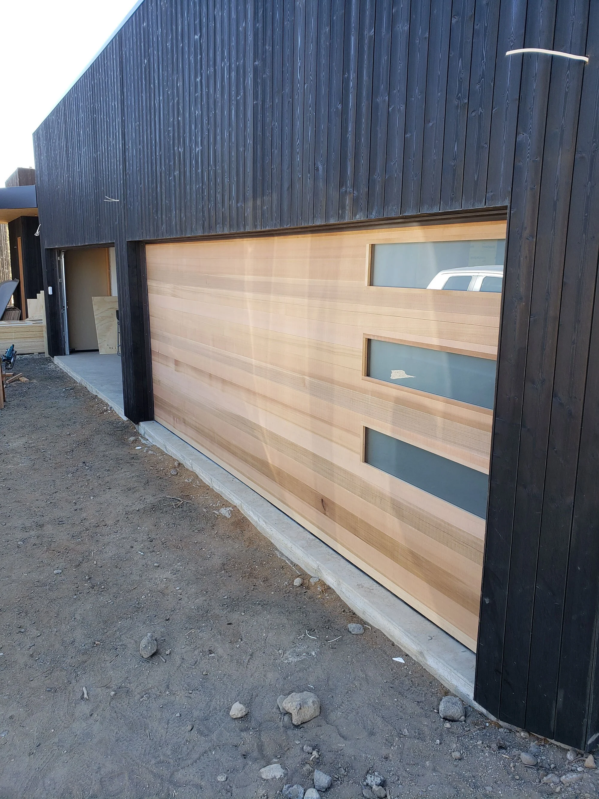 A modern building with a large, wooden horizontal-panel garage door and dark vertical siding, under construction on a dirt lot.