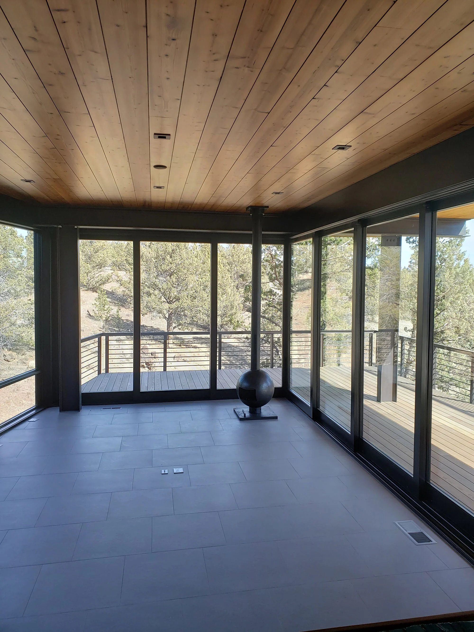 Empty sunroom or enclosed porch with large floor tiles, glass sliding doors, and a wood ceiling, overlooking trees outside.