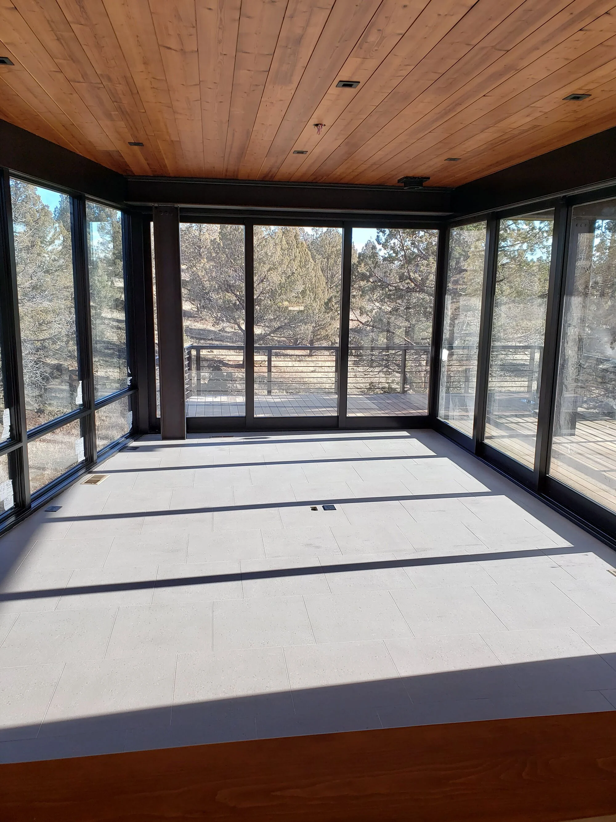Sunlit enclosed balcony with glass walls, wooden ceiling, and tiled floor, overlooking trees and a mountain landscape