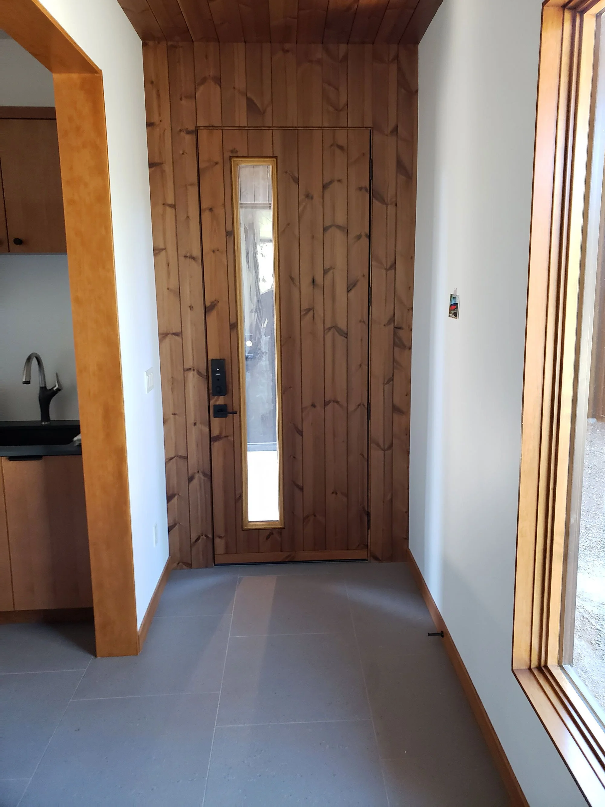 Interior view of a house entryway with wooden panels, a narrow door with a vertical window, and a large window to the side with wooden trim.