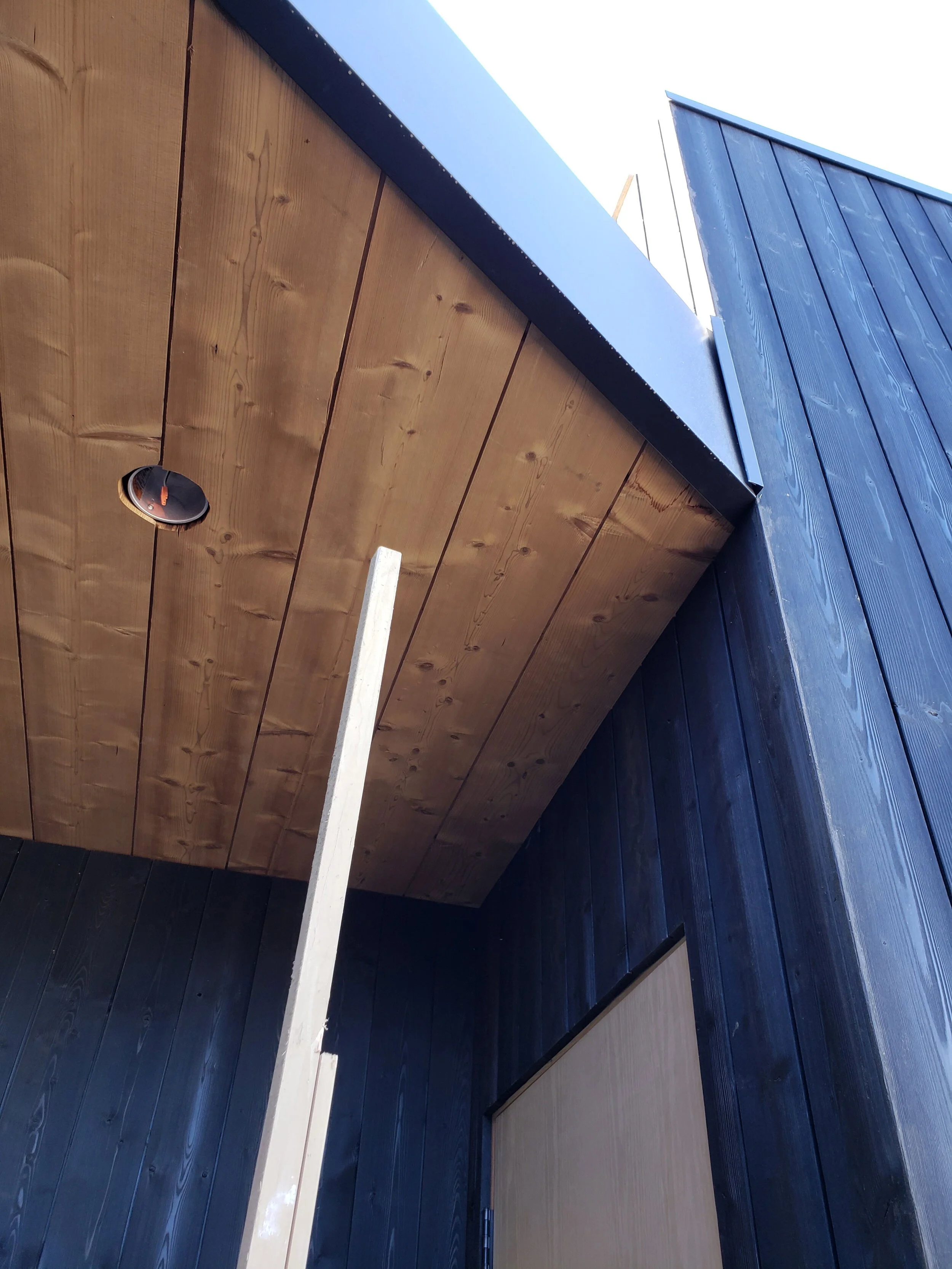 View of building exterior showing black wooden siding, natural wood ceiling, and a wooden post, with an opening in the ceiling for a light fixture.