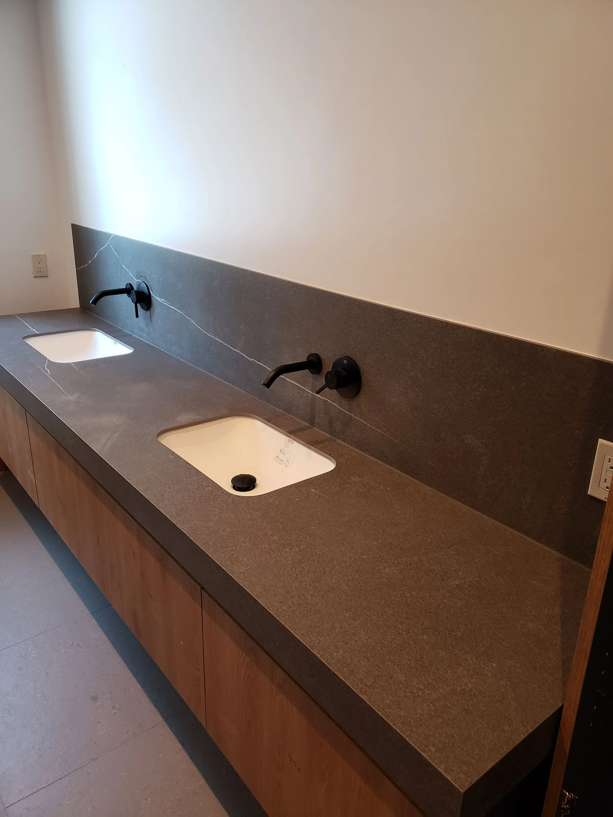 A long brown countertop with two built-in sinks and black faucets, a gray backsplash with white veins, and a power outlet on the right side.