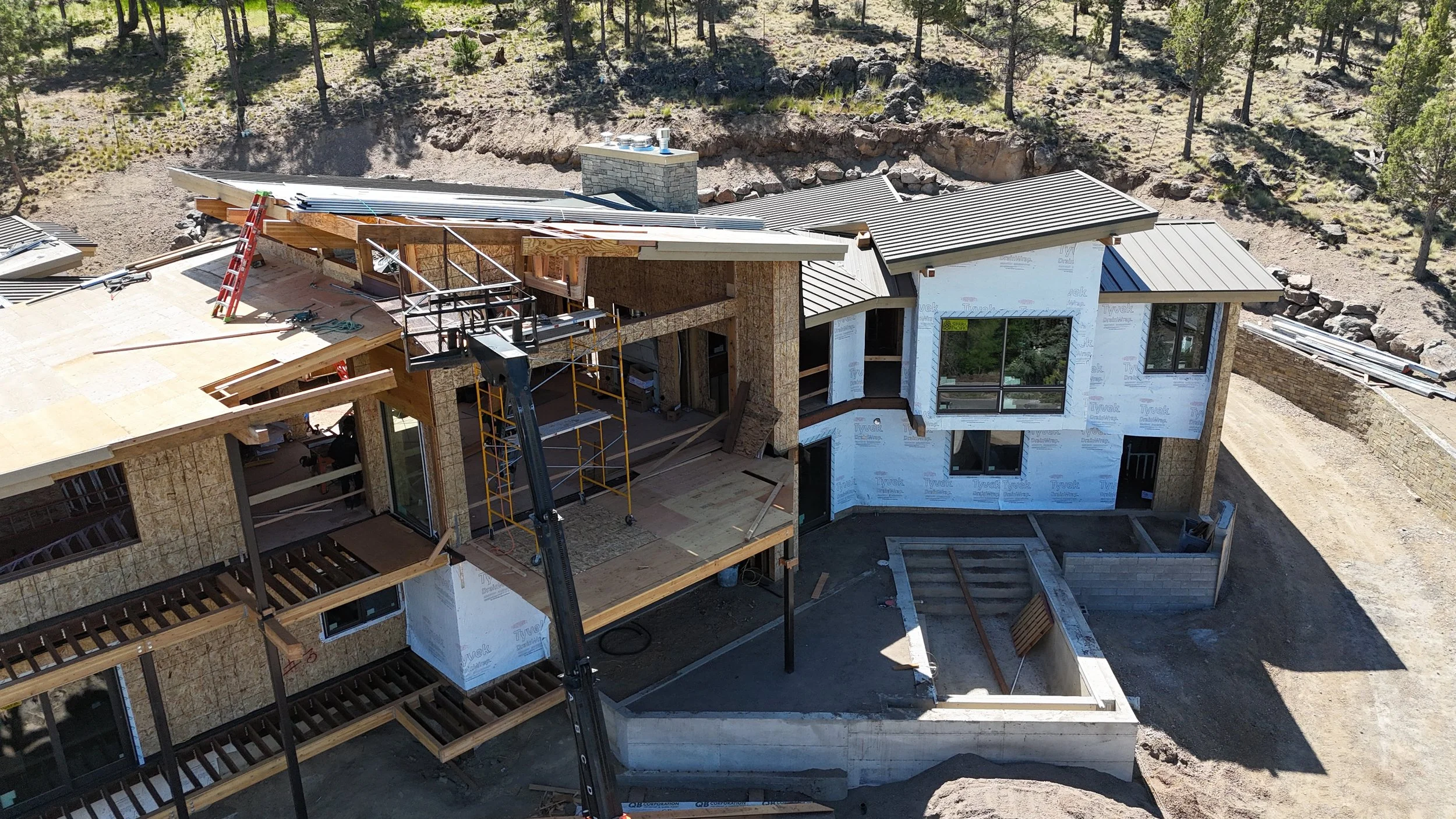 Aerial view of a house under construction with scaffolding, building materials, and partly installed roof, surrounded by trees and rocky terrain.