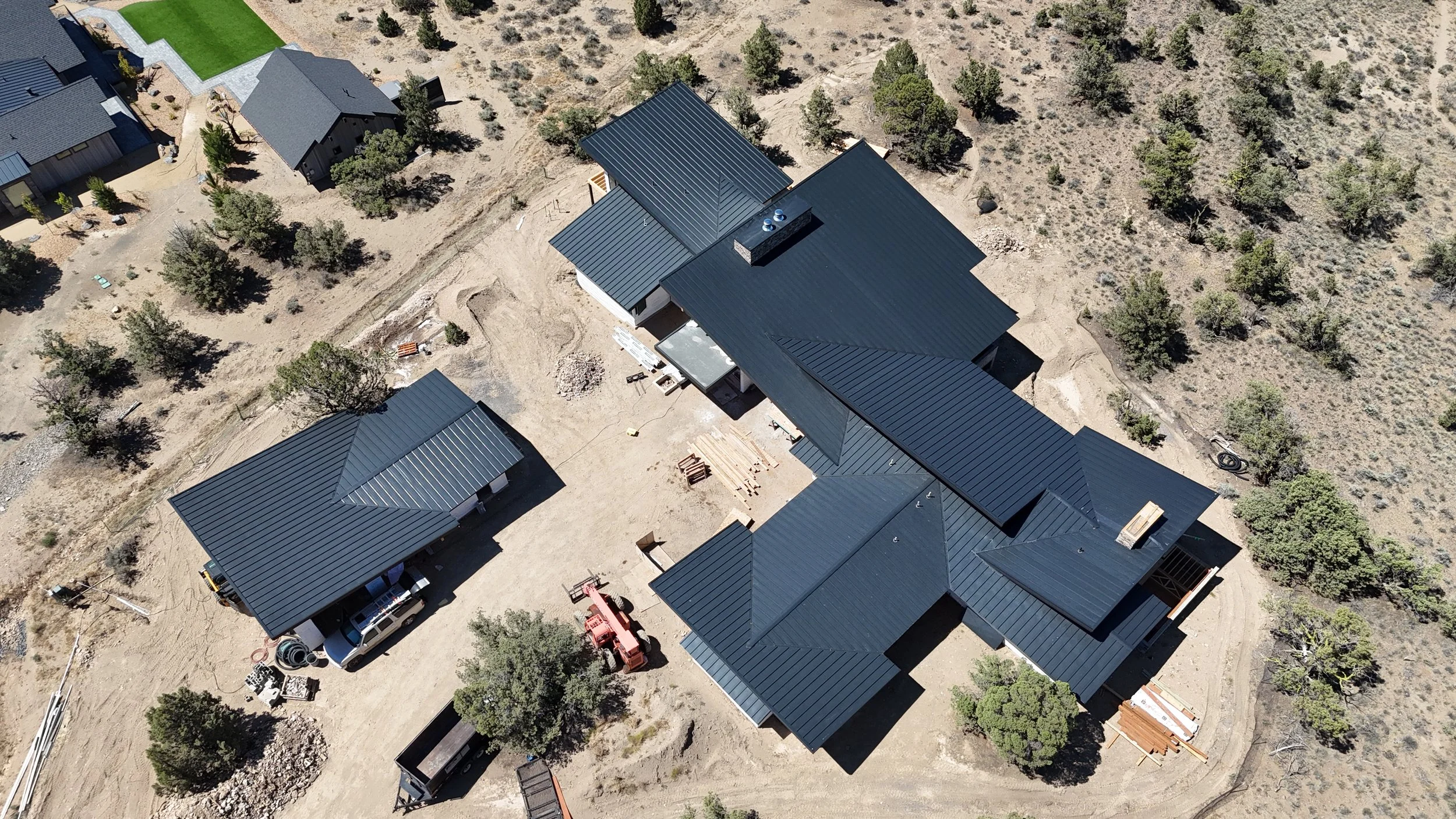 Aerial view of a construction site with multiple houses, black metal roofs, and surrounding trees in a semi-arid landscape.