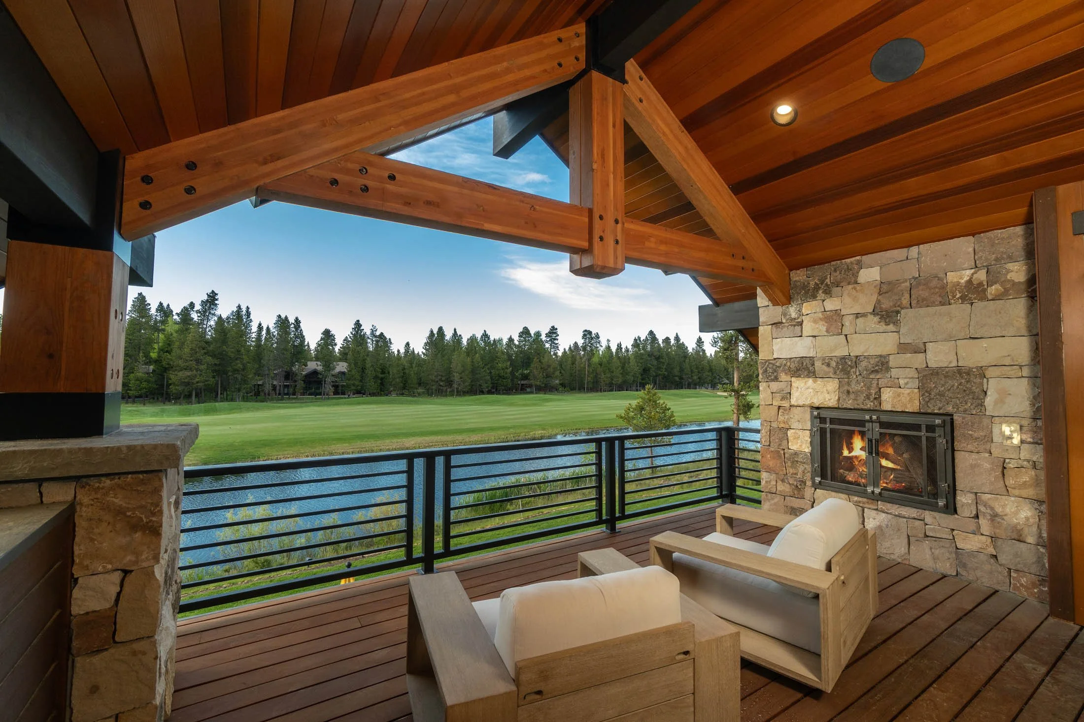 Covered outdoor patio with a stone fireplace and tongue-and-groove wooden ceiling overlooking the water in Crosswater by Pacwest Builders.