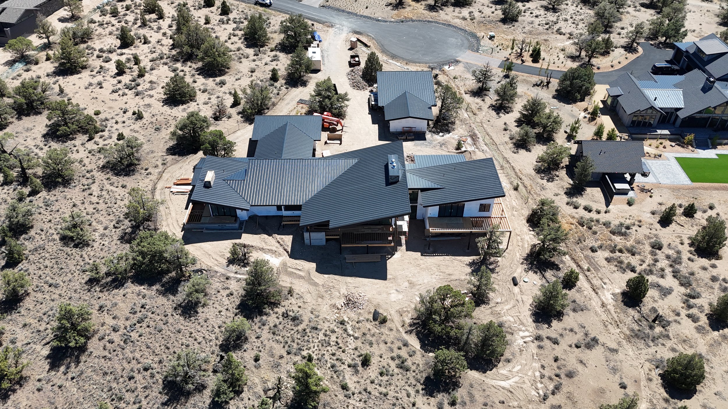 Aerial view of a residential construction site with a large house under construction, surrounded by trees and other completed houses in a semi-arid landscape.