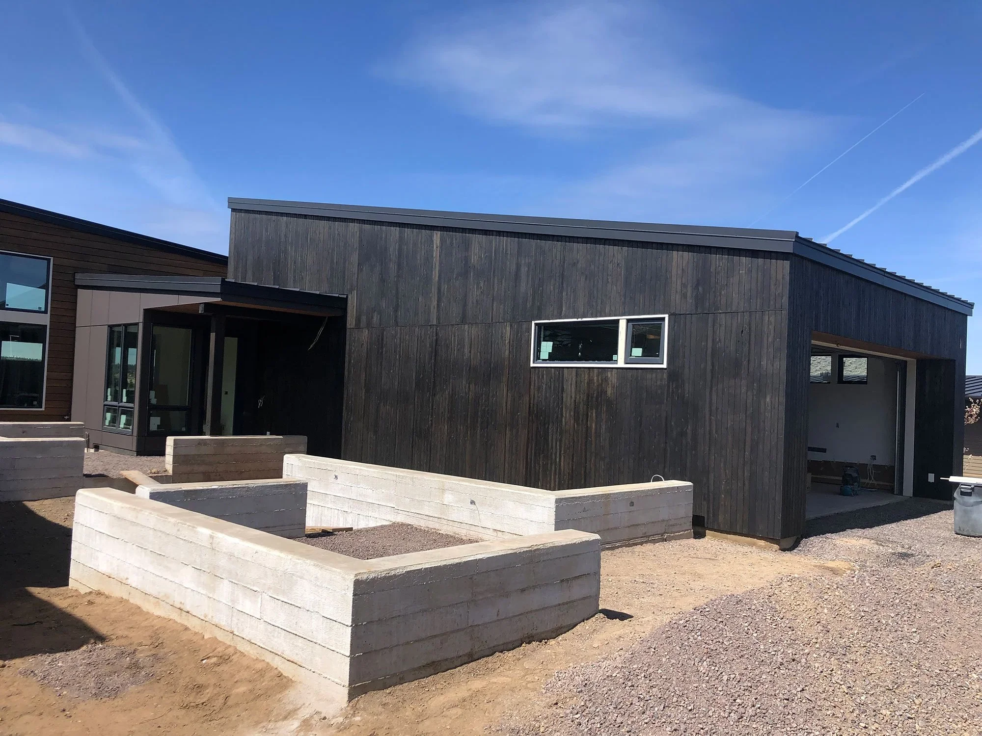 Modern house under construction with black wooden exterior, a smaller rectangular window, and concrete planters in the front yard.