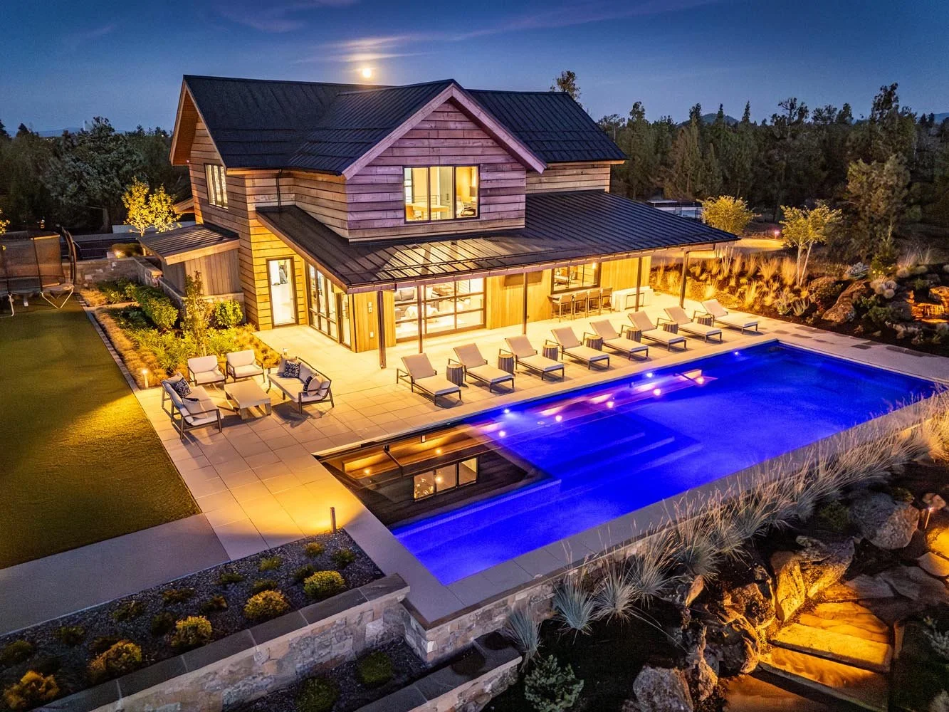 This breathtaking night-time shot captures the essence of luxury living at the Pacific Cascade Heights project, a custom build by Pacwest Builders. The modern guest house, with its striking wood and metal exterior, overlooks a large, inviting illumin