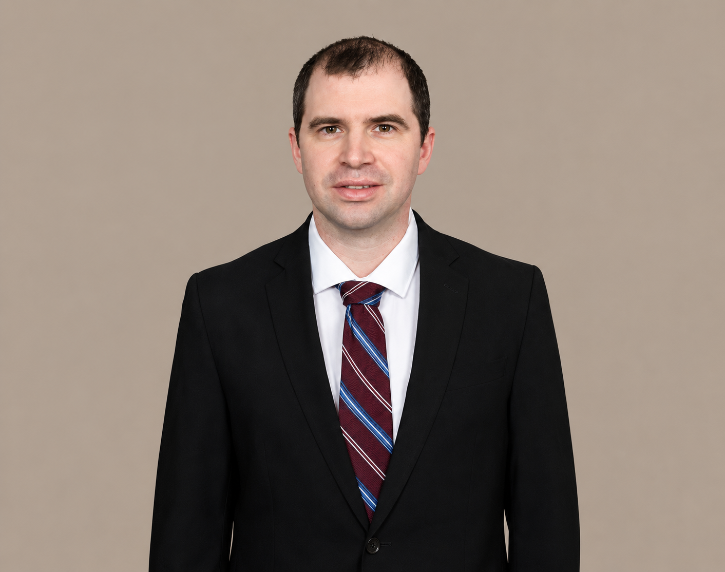 A portrait of a man in a black suit, white shirt, and red striped tie, standing against a beige background.