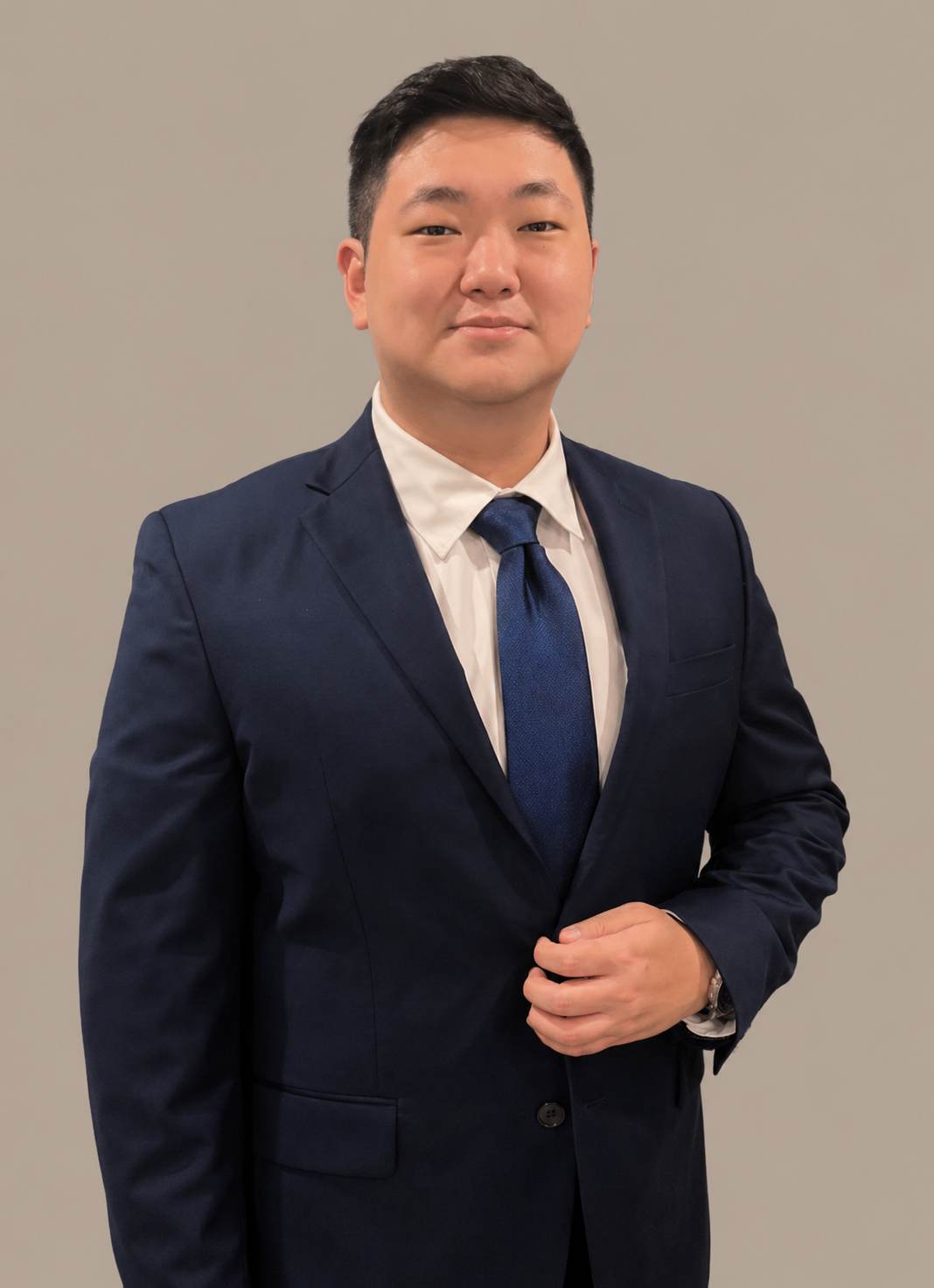 A man in a dark blue suit and tie standing against a plain neutral background.