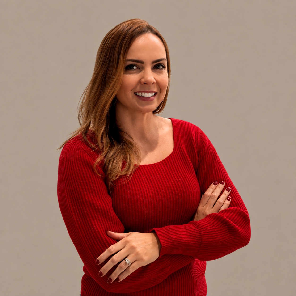 A woman with long auburn hair wearing a red sweater, standing with her arms crossed in front of a plain beige background.
