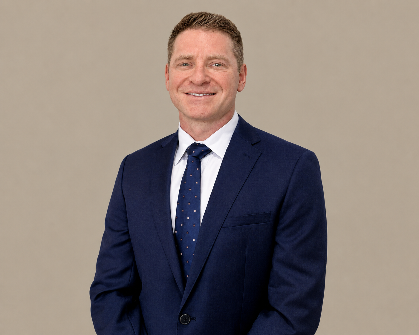 A professional man in a blue suit with a white shirt and blue polka dot tie, smiling, against a plain beige background.