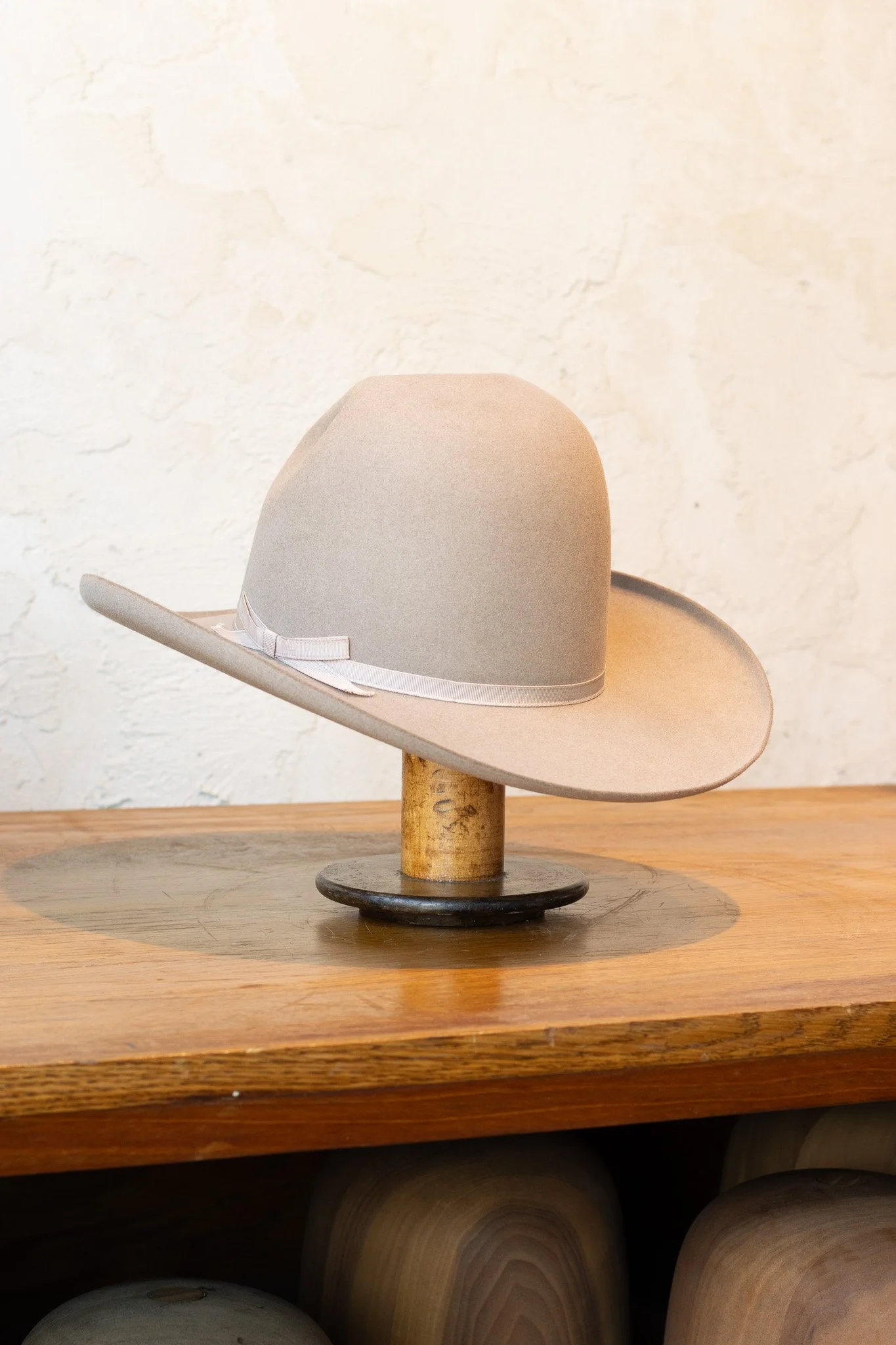 A beige wide-brimmed hat with a white ribbon sits on a wooden table, supported by a wooden stand.