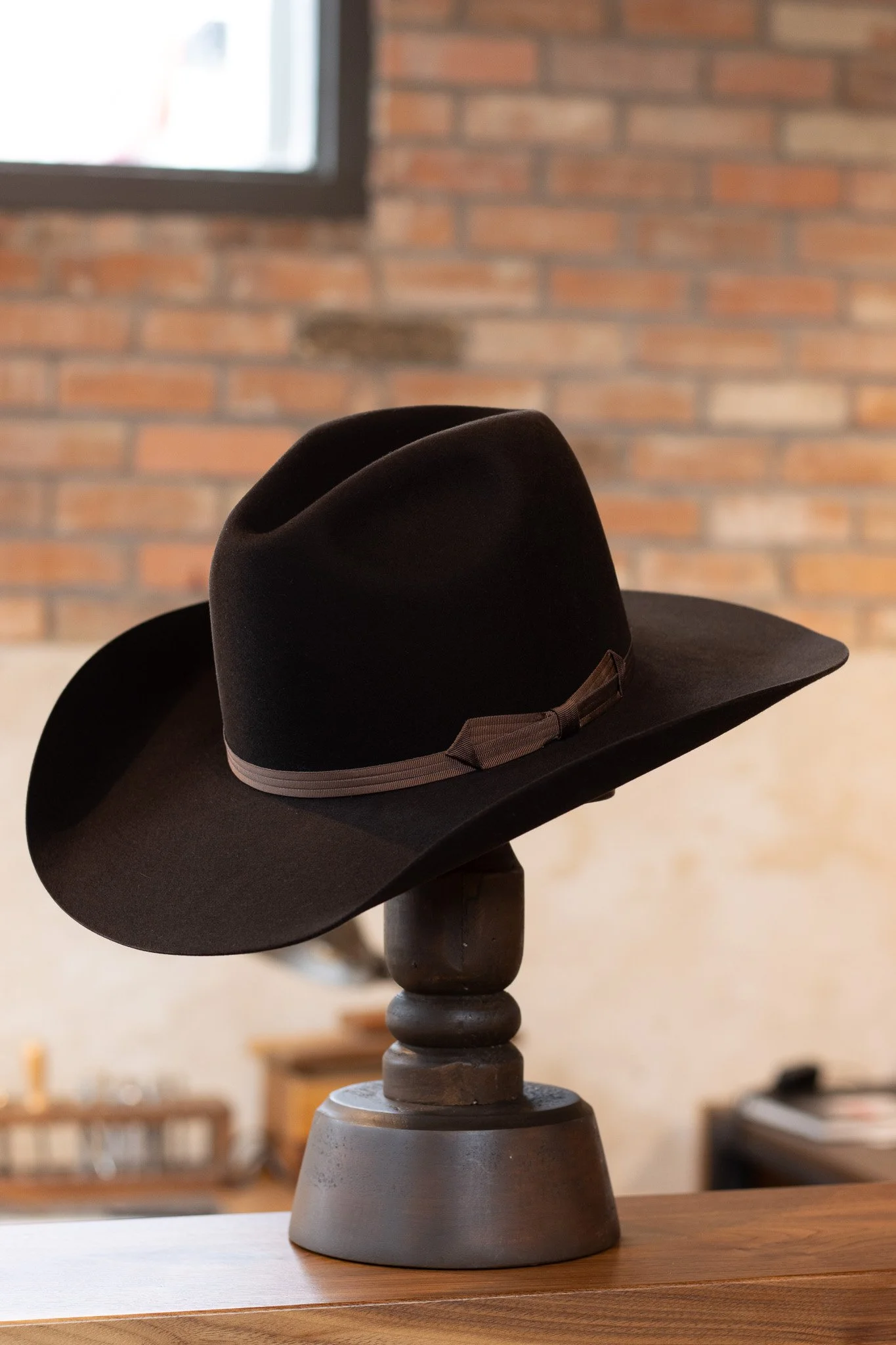 A black wide-brimmed hat with a ribbon and bow, displayed on a dark wooden stand, against a background of brick wall and window.